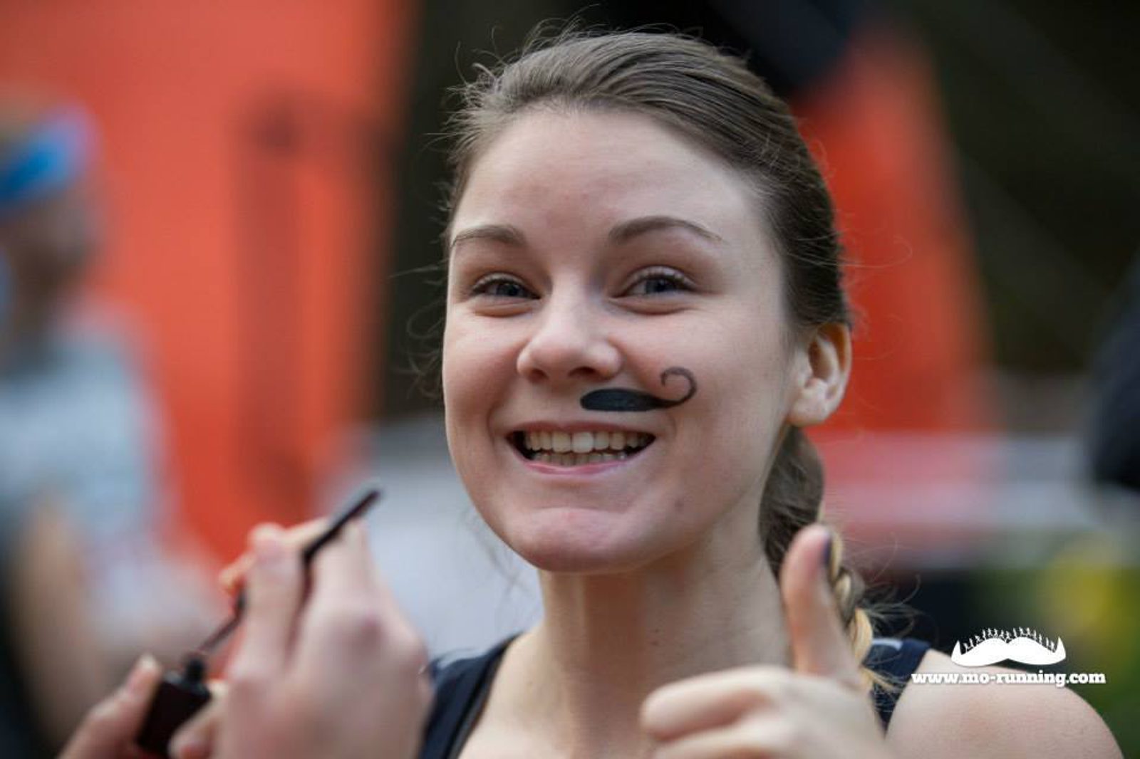 A smiling person with a small painted mustache on their upper lip gives a thumbs-up gesture. The background is blurred, with hints of red and black. The person holds a makeup brush in one hand. The image has the text "www.mo-running.com" at the bottom right.