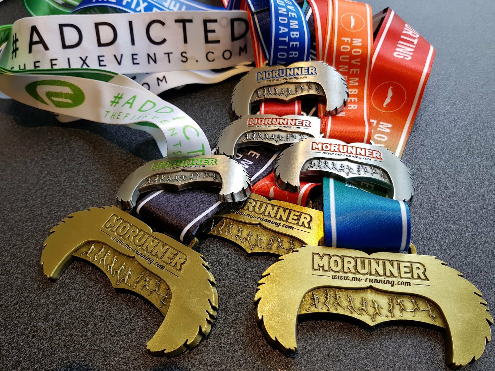 A collection of "MoRunning" race medals shaped like moustaches and a lanyard with various colorful ribbons. The medals feature an imprint of runners on a moustache outline and the text "MoRunner". Nearby is a "#ADDICTED ToTheFixEvents.com" lanyard.