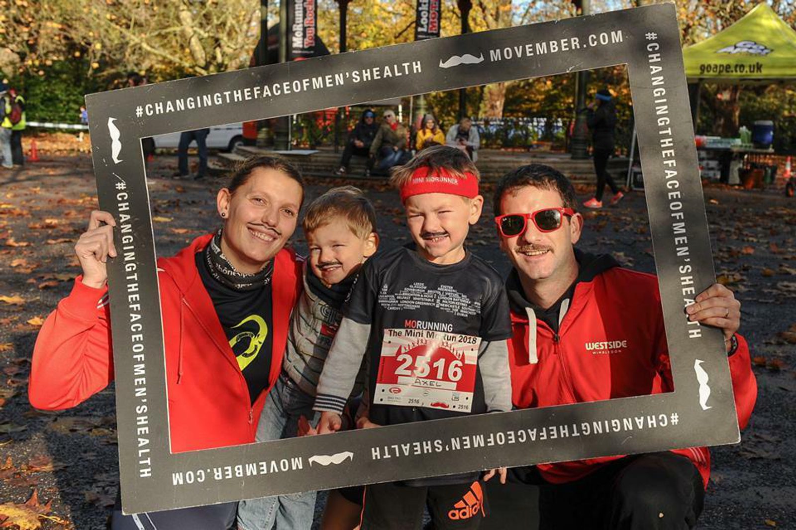 Four people pose for a photo at a Movember event, smiling within a large black frame that reads “#ChangingTheFaceofMen'sHealth.” The child in the center wears a race bib numbered 2516. Fall leaves cover the ground, and other event participants are visible in the background.