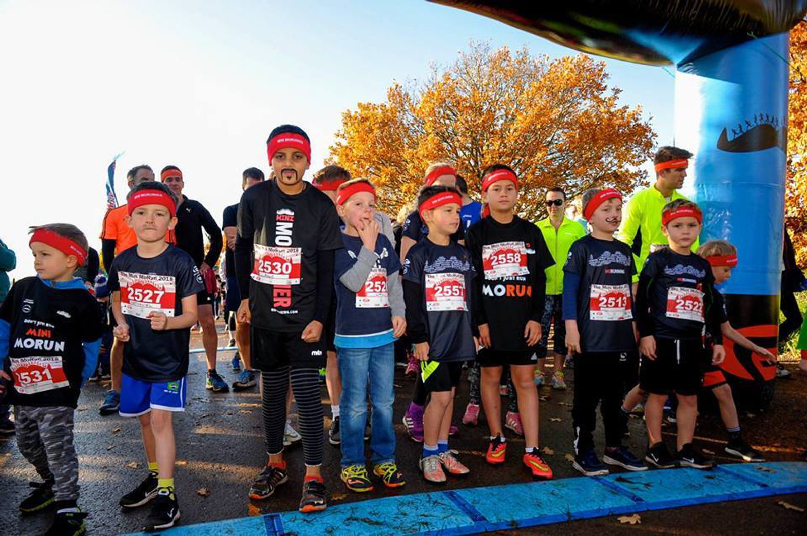 A group of children, wearing black and red headbands, stand at the starting line of a race outdoors on a sunny day. They are accompanied by a few adults. Trees with autumn foliage are visible in the background. Most participants are wearing race bibs with numbers.