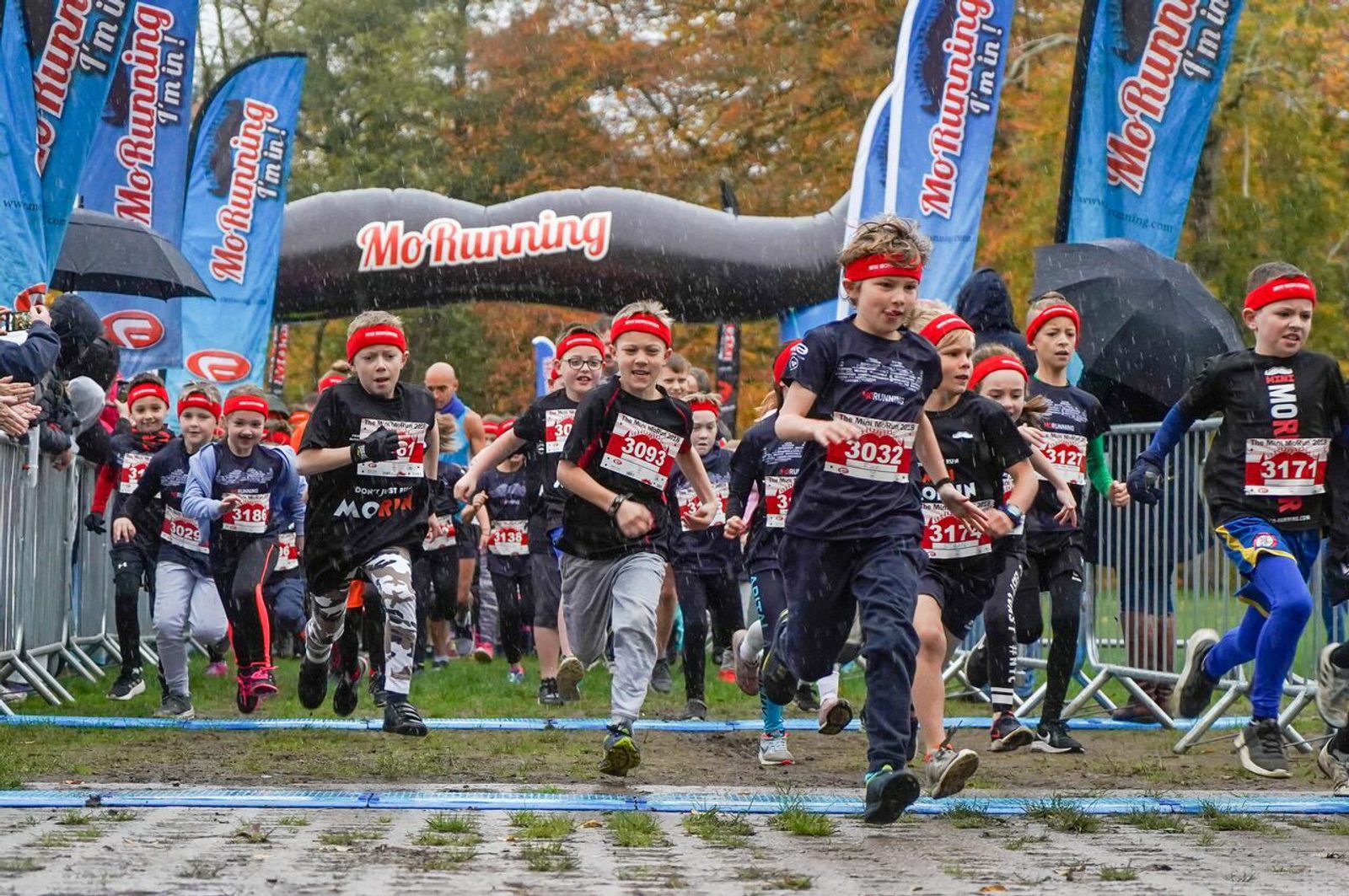 A group of children in athletic attire, most wearing red headbands, are running at the beginning of a race marked with a "MoRunning" inflatable arch. Spectators hold umbrellas in the background as the race takes place on a wet, overcast day.