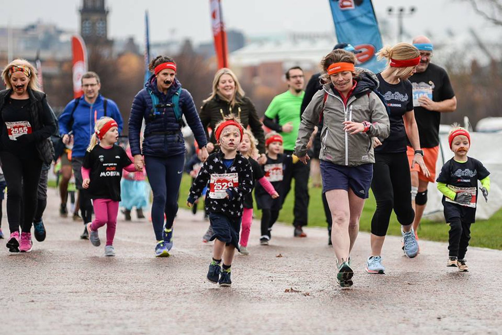 A diverse group of people, including children and adults, are running and walking in an outdoor event. Participants wear numbered bibs and some have red headbands. The background shows a cloudy sky and urban buildings. Everyone appears to be enjoying the activity.