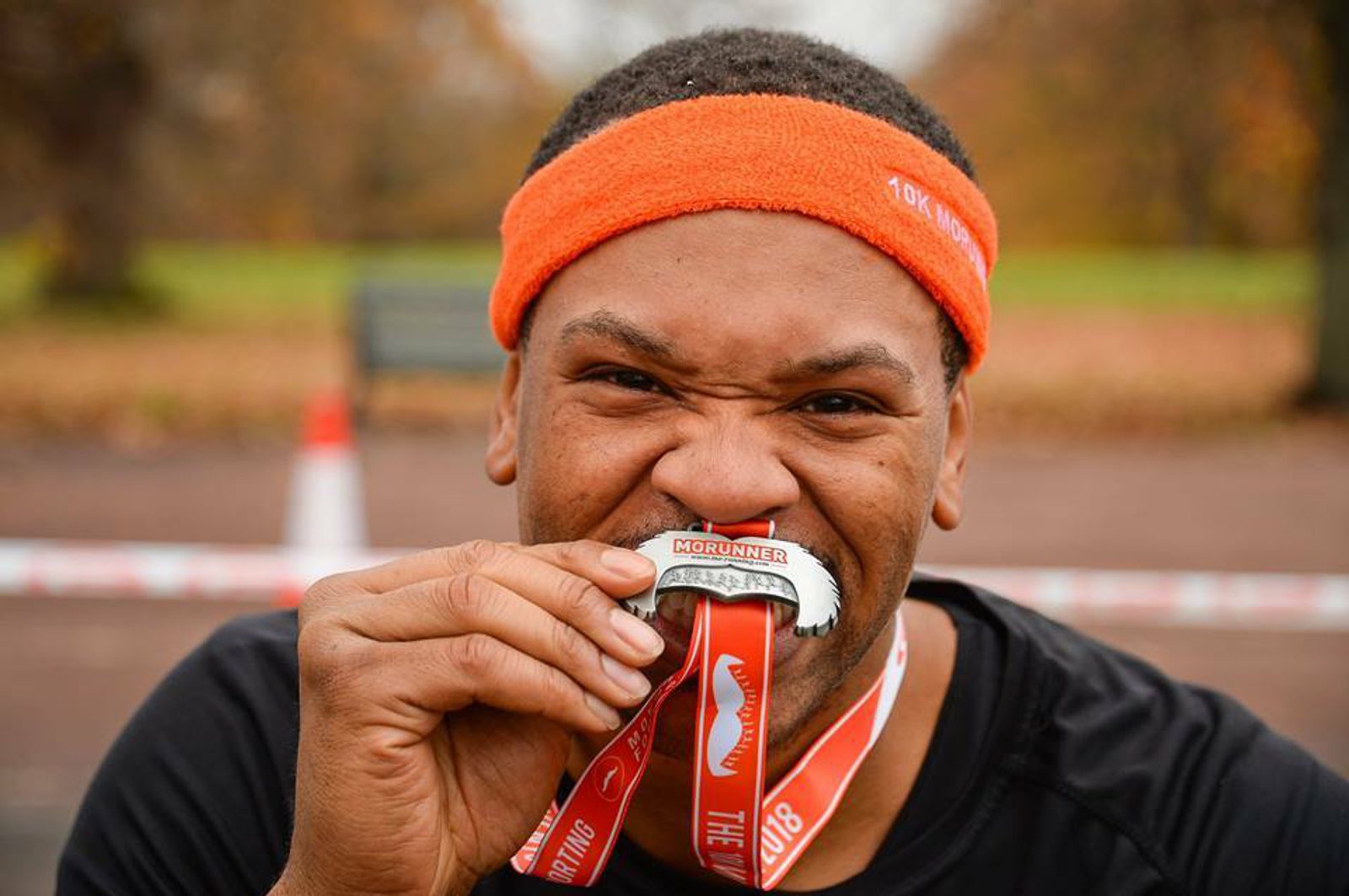 A man wearing an orange headband and a black shirt bites a silver medal ribboned with red lanyard. He looks energetic and appears to be at an outdoor event, possibly concluding a race or sporting event. The blurred background shows autumn foliage and a distant bench.