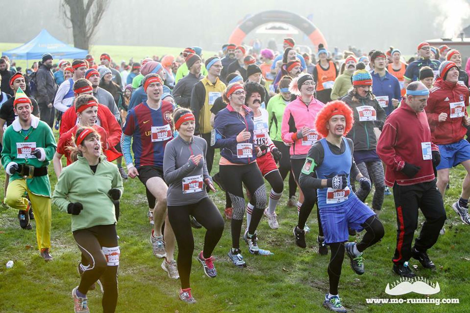 A group of runners in colorful outfits and costumes participate in a fun run. Many are wearing bright clothes, and some have wigs or themed hats. The event appears lively and spirited, taking place outdoors with trees and grass visible in the background.