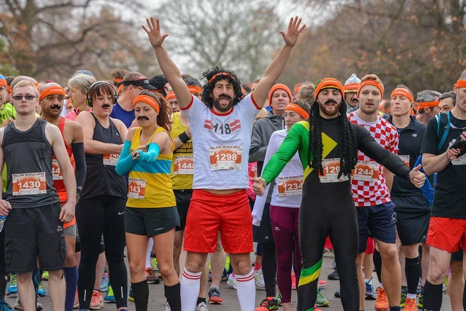 A group of people at the start line of a race, many wearing colorful costumes and orange headbands. The front center runner, dressed in a 70s athletic outfit with a sticker reading "1298," has raised arms, and others around him exhibit various humorous outfits and poses.
