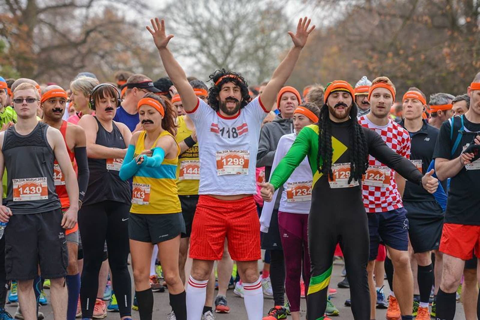 A group of runners in colorful and whimsical outfits are gathered at the starting line of a race. Some are wearing orange headbands and fake mustaches. Numbered race bibs are pinned to their clothing, and one runner, wearing a white shirt, is raising his arms.