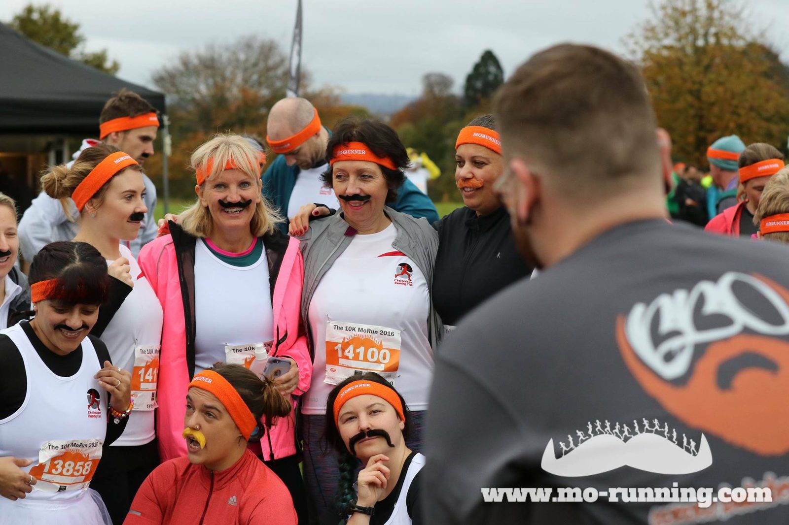 A group of people wearing orange headbands and fake moustaches pose for a photo before participating in a fun run event. One individual in the foreground, wearing a shirt with the "Mo-Running" logo, faces the camera while others smile and laugh.