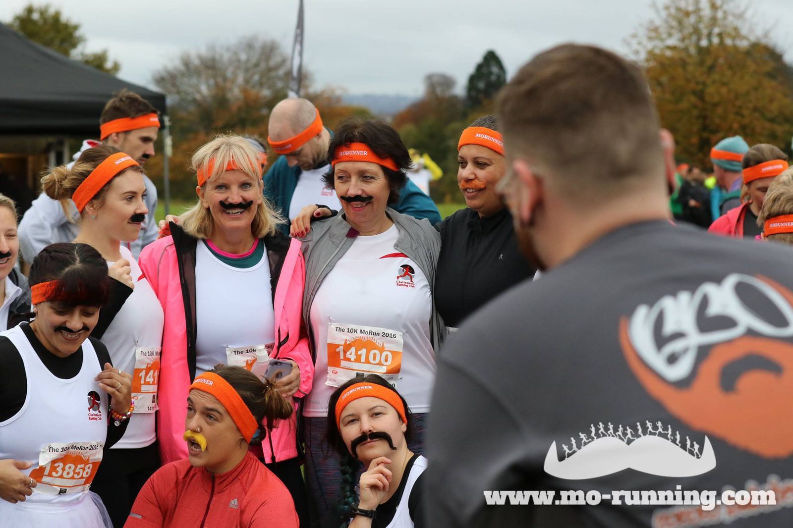 A group of people wearing white shirts and orange headbands gather for a photo at a fun run event. Many of them are wearing fake mustaches, and some are playfully posing with yellow whistles in their mouths. In the foreground, a man in a gray shirt is facing away.