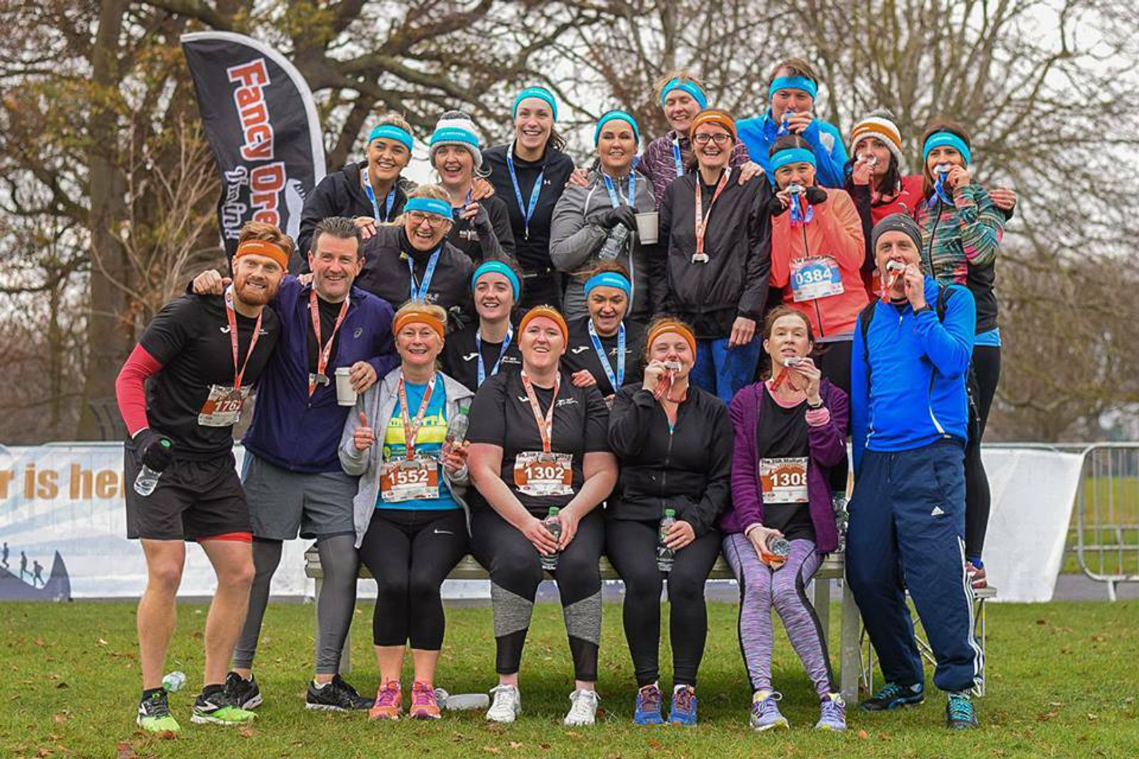 A group of runners pose proudly for a photo after a race, wearing colorful headbands and medals around their necks. They are standing and kneeling on grass with a banner and trees in the background. Some runners are playfully biting their medals.