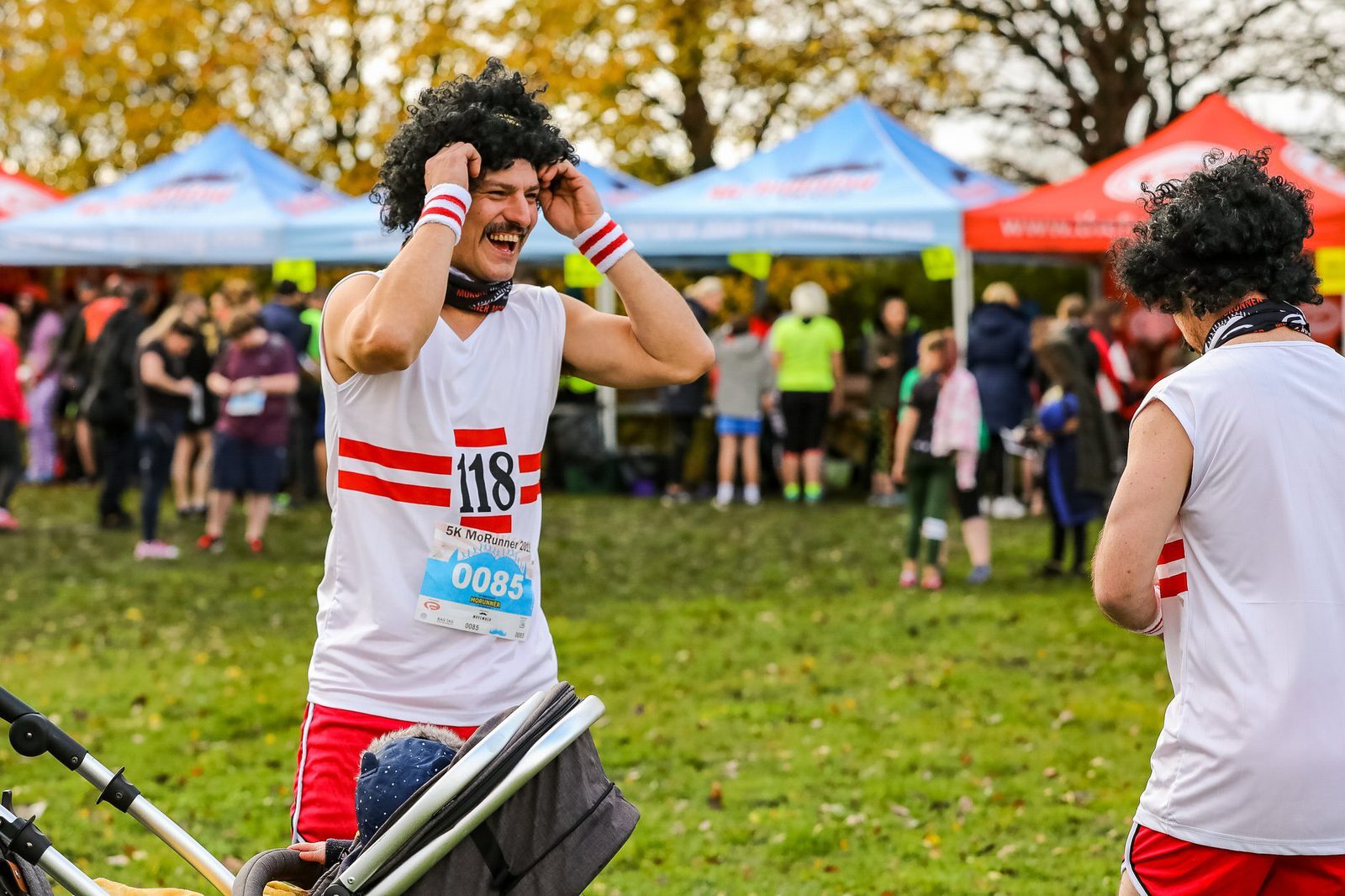 Two people, dressed in matching red and white athletic costumes, adjust their curly black wigs at an outdoor event. One person's race bib reads "118" and the other's reads "085." A group of spectators and blue and red tents are visible in the background.