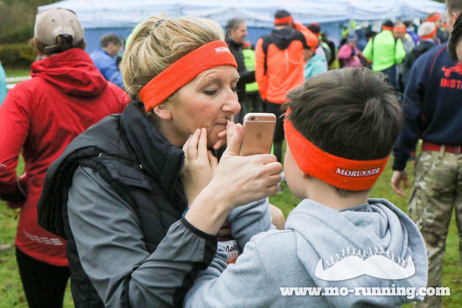 A woman and a young boy, both wearing orange headbands, stand together at an outdoor event. The woman is holding a phone in front of her face, and the boy is helping her adjust or check her appearance. Other participants and tents are visible in the background.