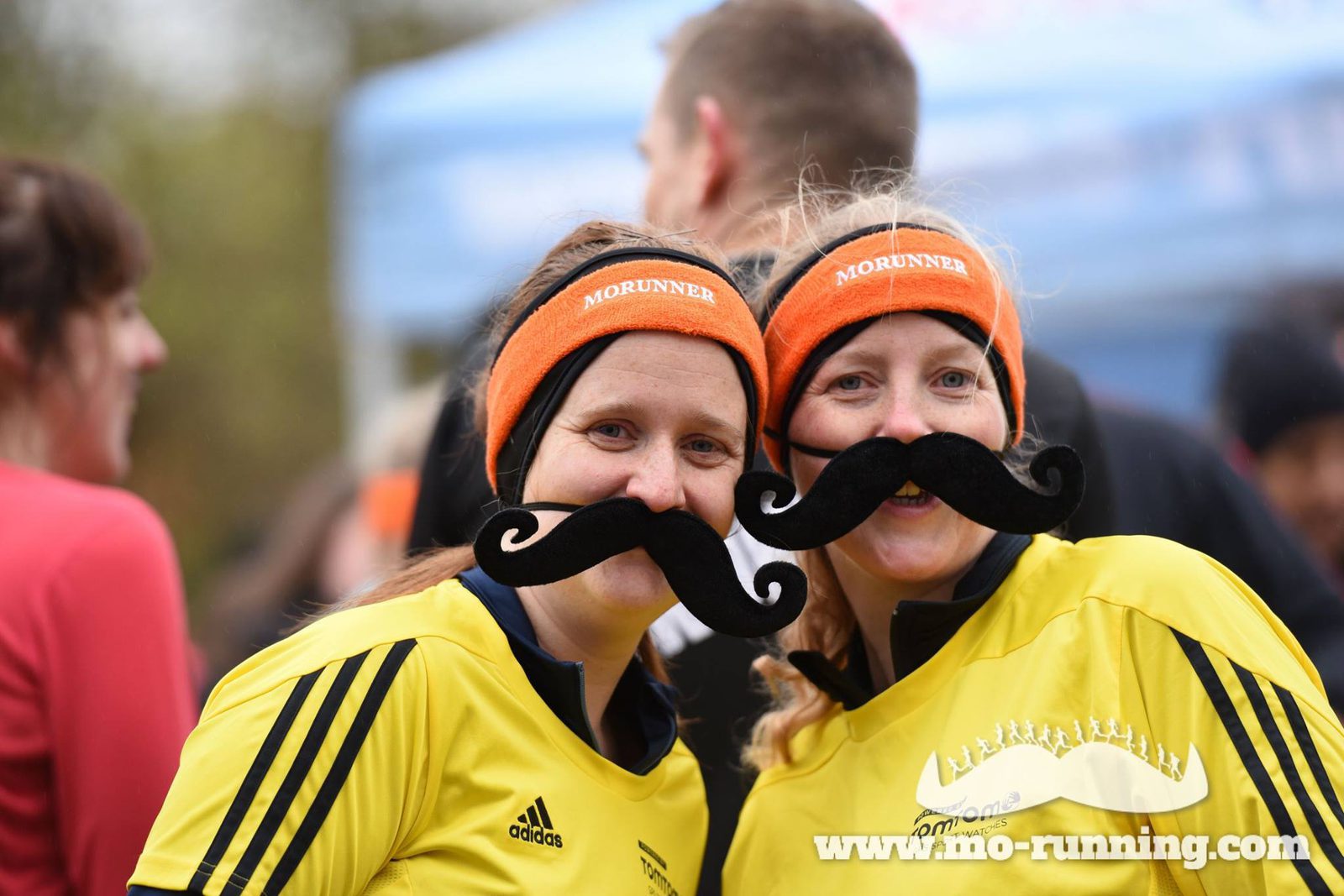 Two smiling women wearing matching yellow athletic shirts, orange headbands, and fake black mustaches stand close together. They are part of a running event, evidenced by the banner and people in the background. The website "www.mo-running.com" is visible at the bottom.