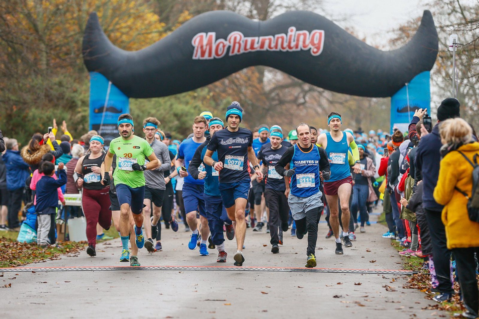 A large group of runners, many wearing blue headbands, participate in a race. An inflatable arch with a large mustache and the text "Mo Running" marks the start/finish line. Spectators, some taking photos, watch from the sides on a tree-lined pathway.