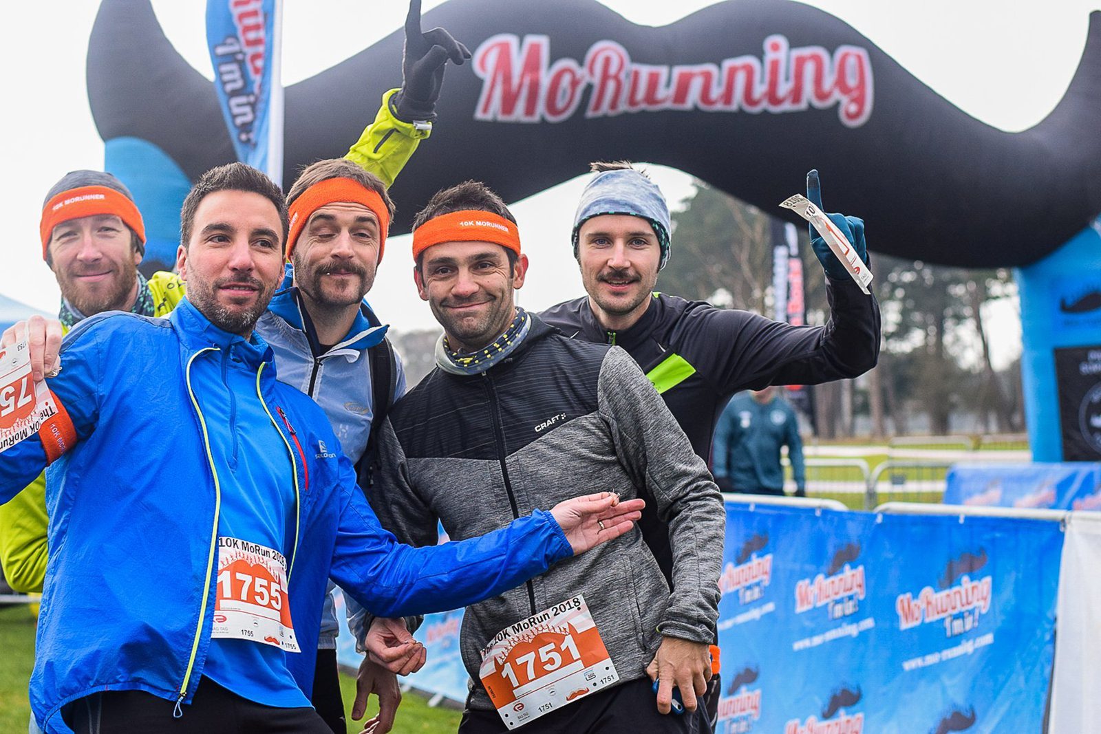 Five men wearing running gear and race bibs pose for a group photo at a MoRunning event. The archway behind them is decorated with a large mustache, and the participants are smiling and making celebratory gestures.