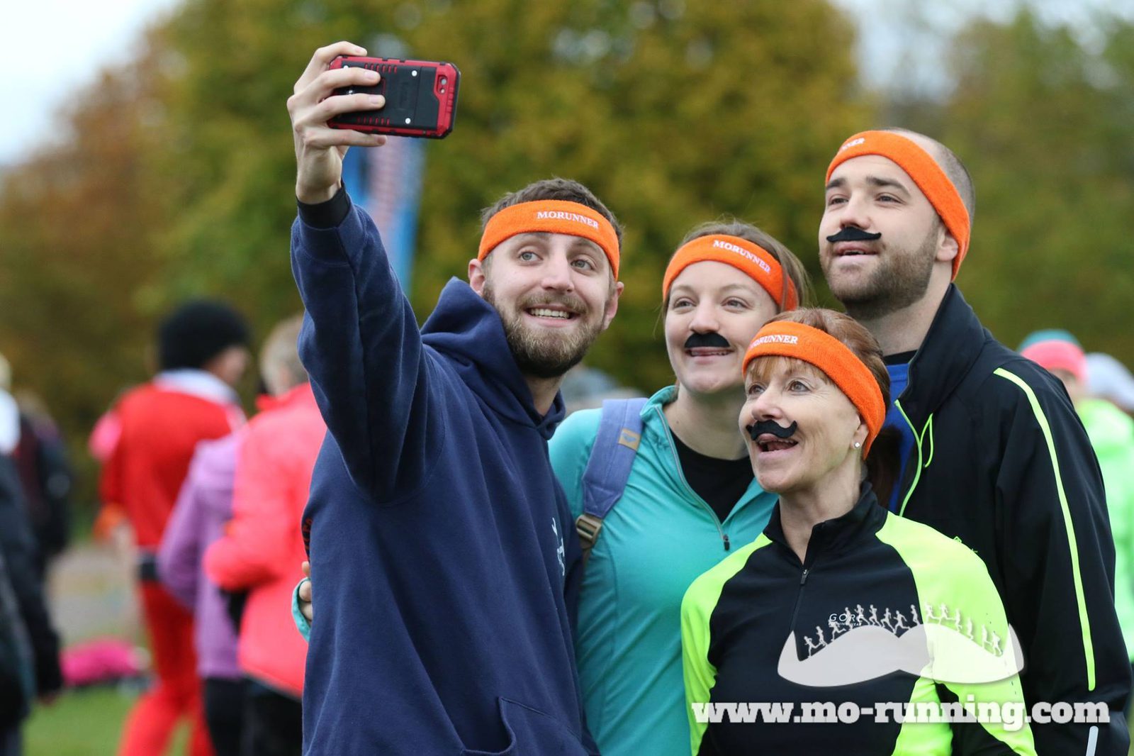 A group of four people, with faux mustaches and orange headbands, take a selfie at an outdoor event. The headbands read "MO". They are dressed in colorful athletic clothing with trees in the background. The event website "mo-running.com" is visible in the corner.