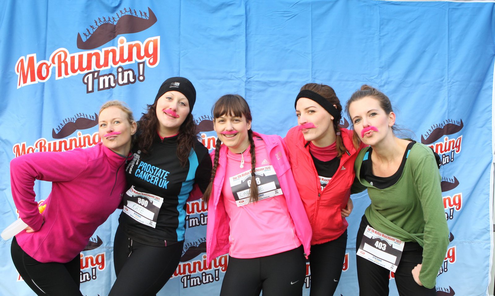 A group of five women with fake mustaches pose in front of a "Mo Running I'm in!" banner. They are dressed in athletic clothing, each wearing a bib number, suggesting they are participating in a running event, likely in support of prostate cancer awareness.