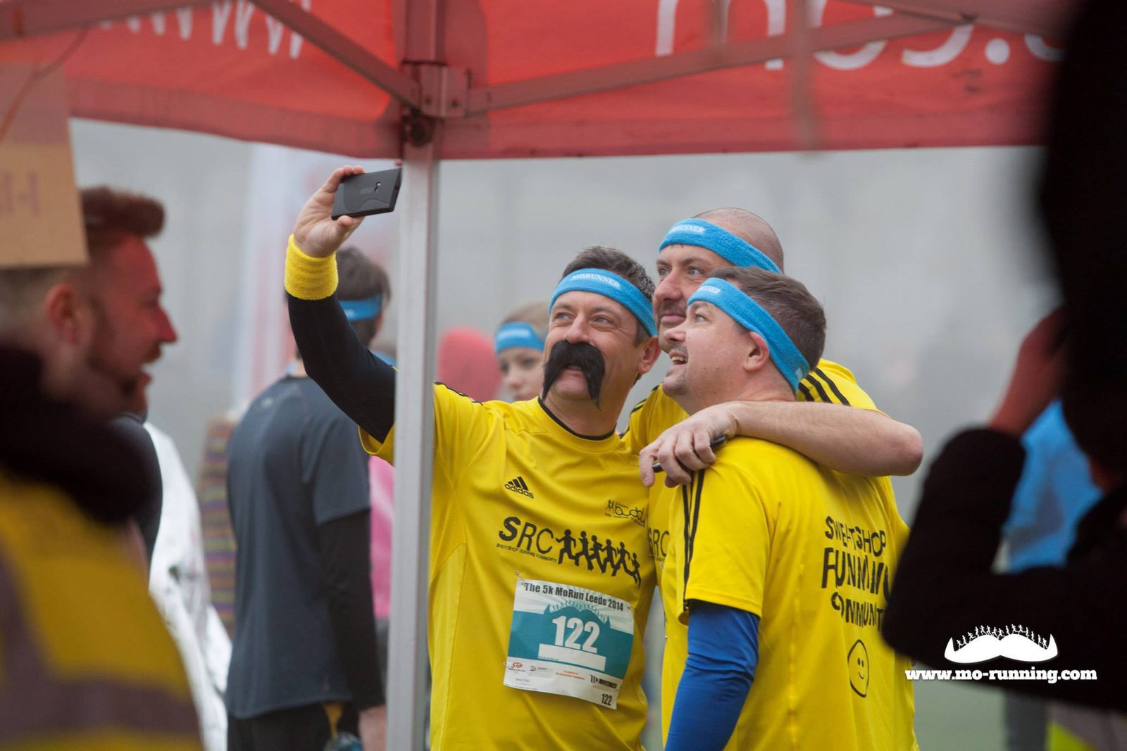 Three men wearing yellow shirts and blue headbands with fake mustaches pose for a selfie under a red canopy at a running event. One man holds the phone, while the others smile and lean in. The event logo "mo-running" is visible on the canopy.
