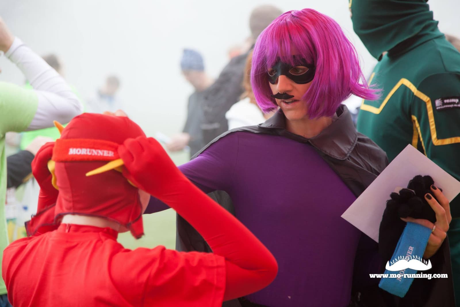 Two people in colorful costumes engage in conversation at an outdoor event on a foggy day. One, dressed in red with yellow ears, adjusts their headgear, while the other, in a purple wig and mustache, holds documents. The atmosphere is festive and lively.