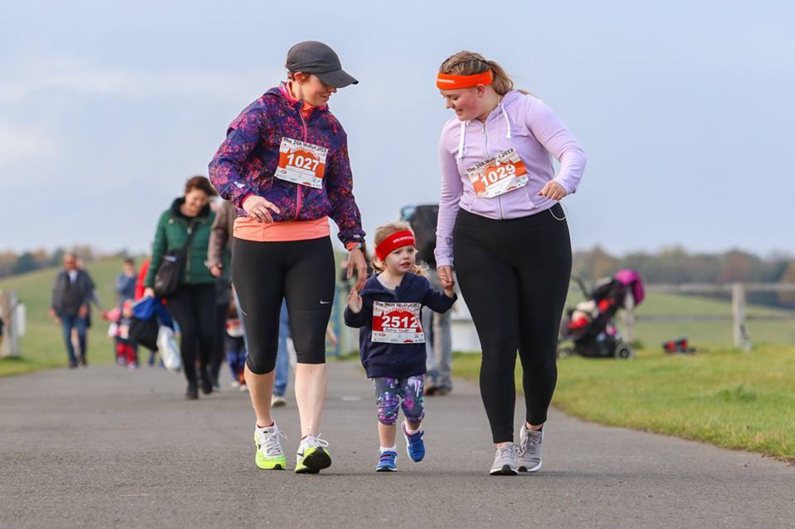 Two women and a child are participating in a race. The women, wearing athletic gear, are each holding one of the child's hands as they walk down a paved path. Other participants and spectators are visible in the background. The atmosphere is lively and supportive.