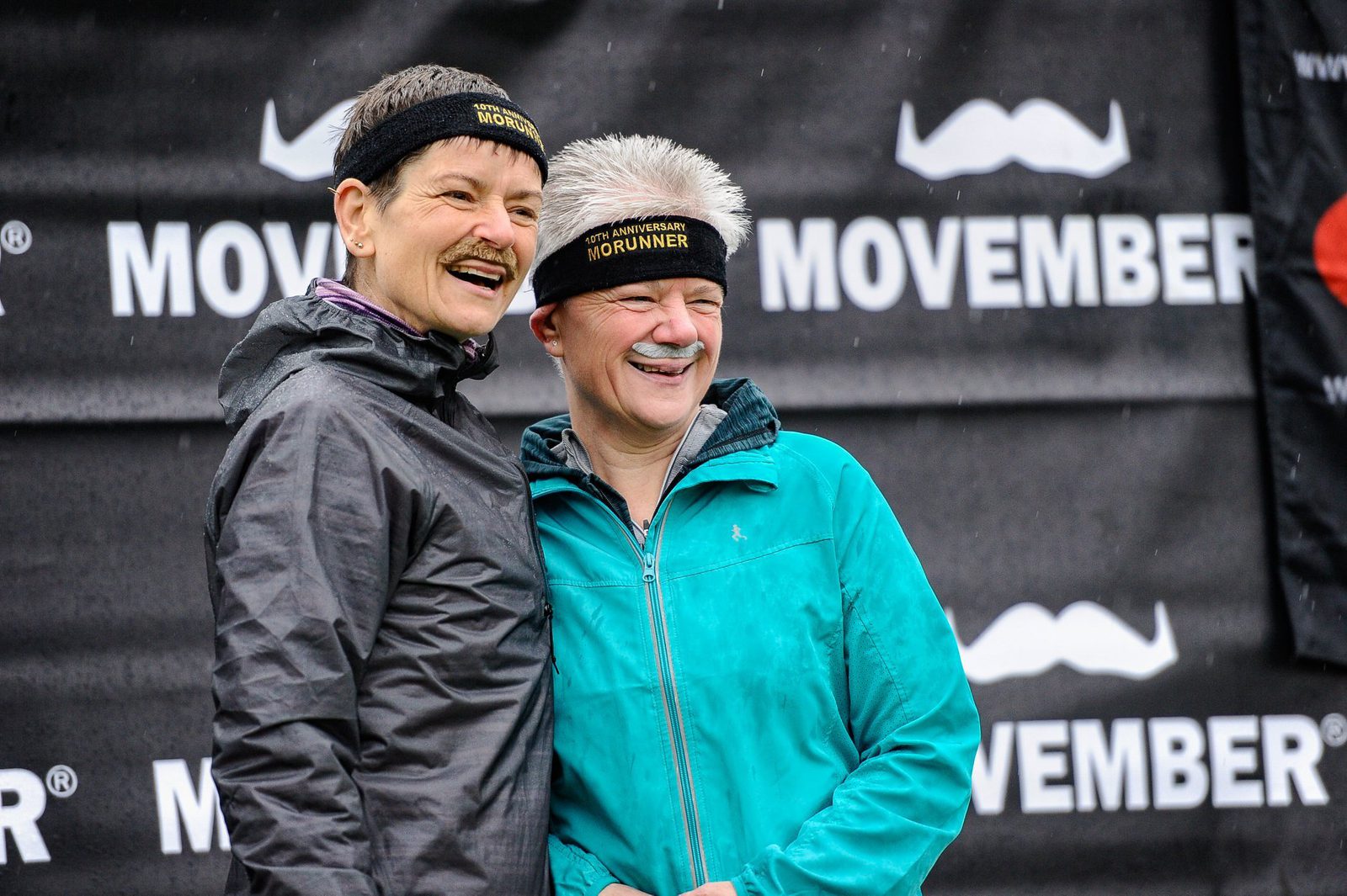 Two people wearing headbands that read "MOURUNNER" stand close together, smiling. Both sporting fake mustaches, they are in front of a backdrop featuring "MOVEMBER" and mustache icons, seemingly participating in an event promoting the Movember cause.