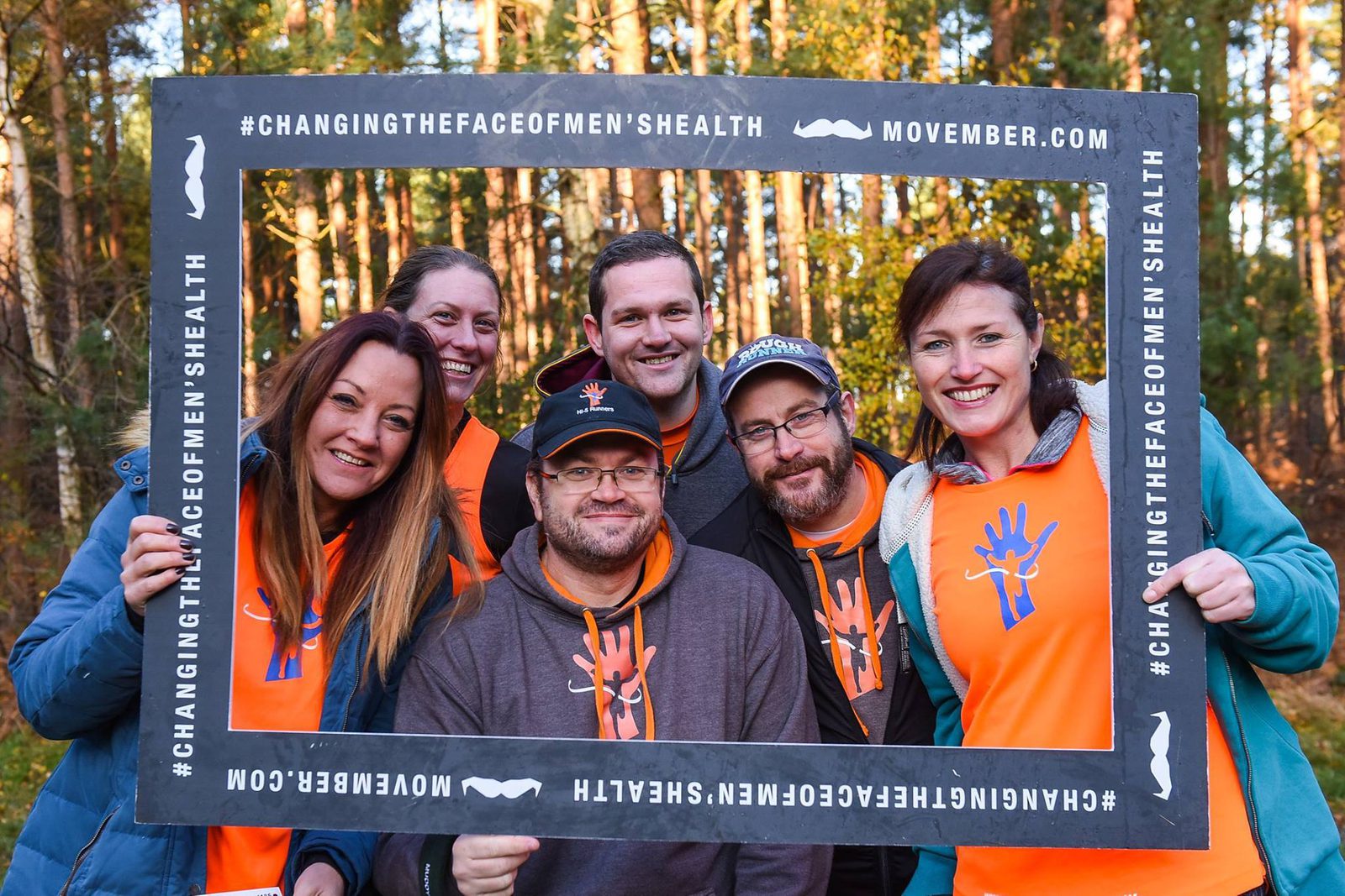 A group of six people, three in the back and three in the front, smile while standing outdoors. They are holding a large picture frame with the text "#ChangingTheFaceOfMensHealth" and "Movember.com" on the borders. Trees are visible in the background.