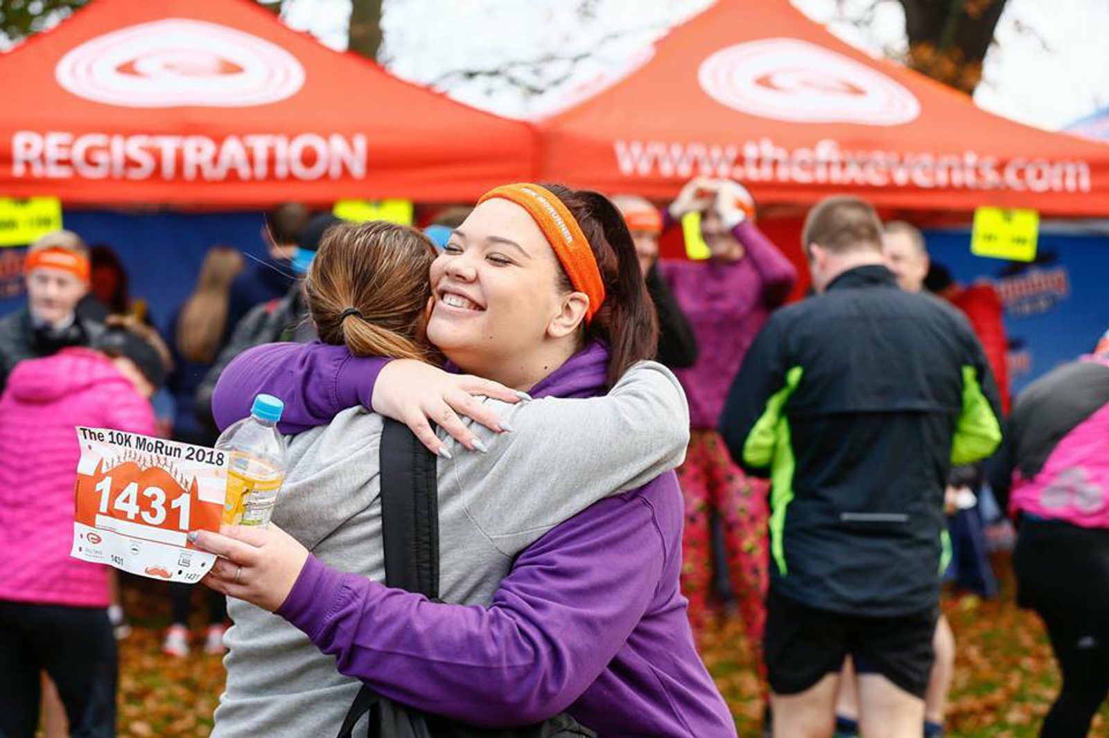 Two women are embracing after participating in a race. One woman, facing the camera, is holding a race bib numbered 1431 and a bottle of water. Behind them is a red registration tent with more race participants milling around. The mood is celebratory.