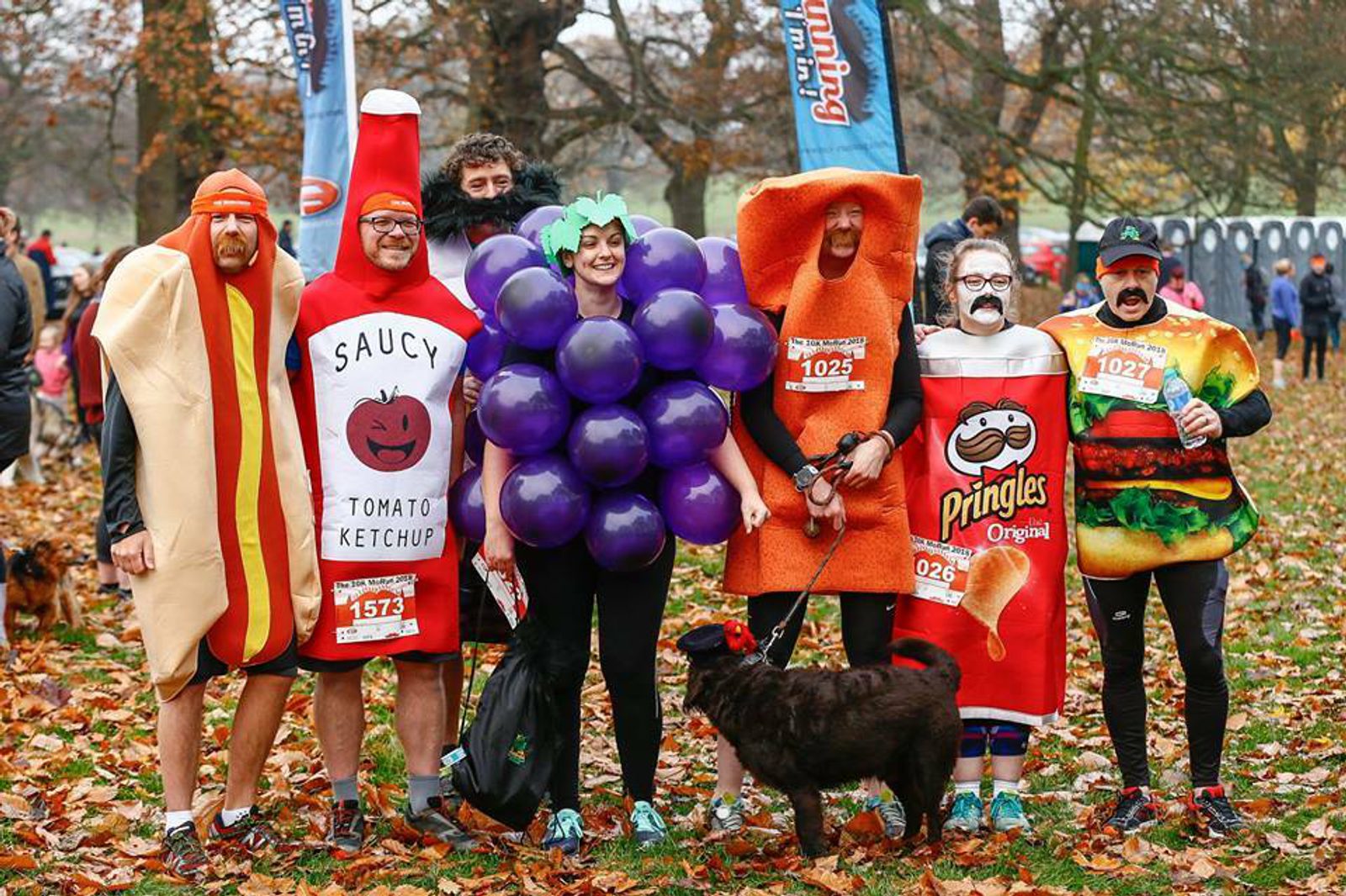 A group of people dressed in various food costumes, such as a hot dog, ketchup bottle, bunch of grapes, and Pringles can, pose together in a park with autumn leaves on the ground. There is also a dog standing near the group. They seem to be participating in an event.