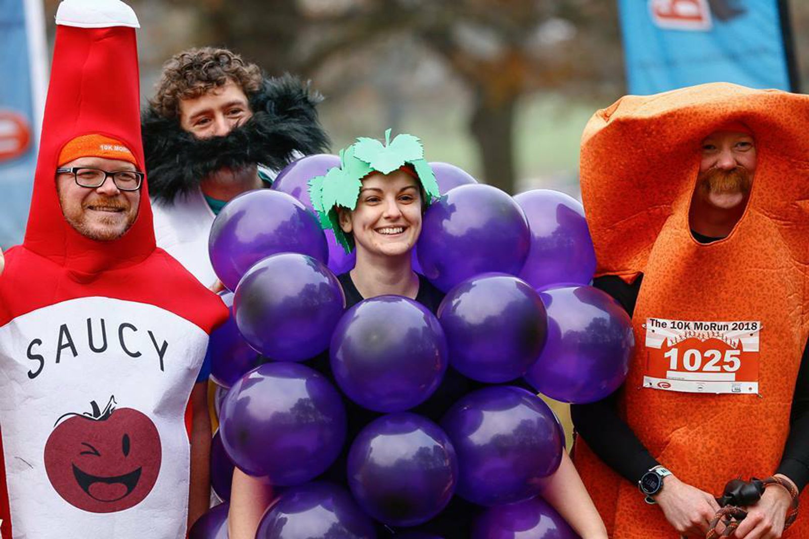 Four people are in costume posing for a photo. From left to right: a person dressed as a sauce bottle, a person with a black beard, a person dressed as a bunch of grapes, and a person in an orange costume with a race bib for "The 10K MoRun 2018" numbered 1025.