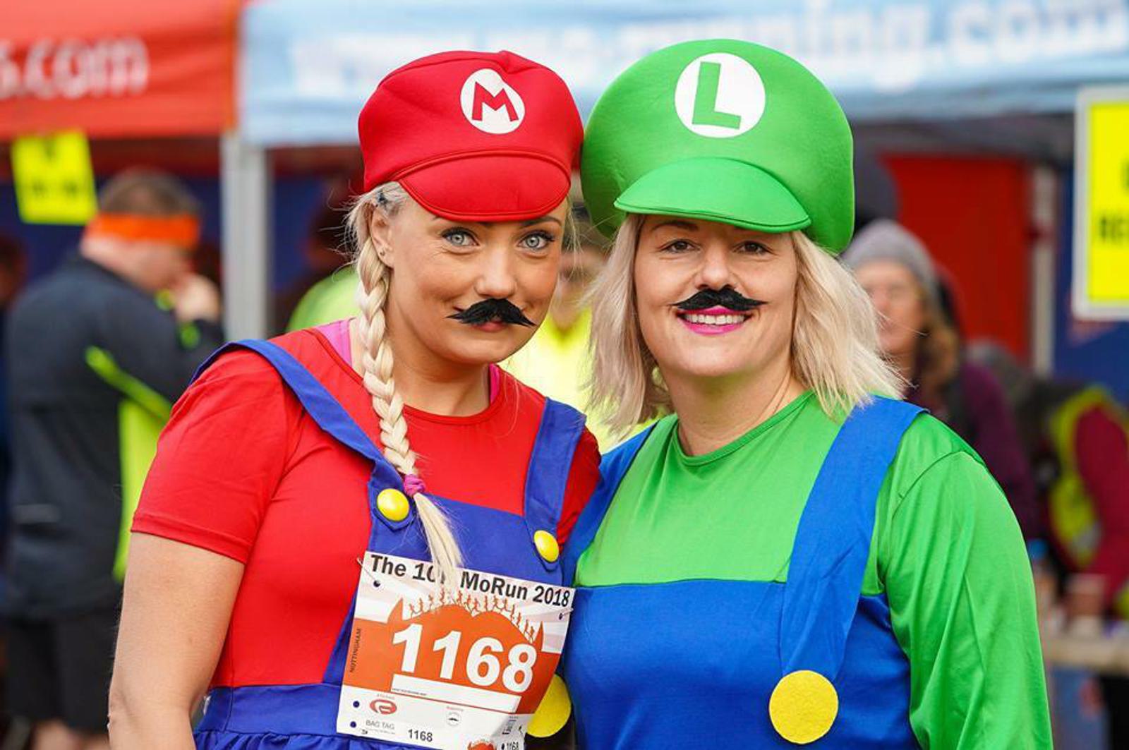 Two people dressed as popular video game characters stand together, smiling at a race event. The person on the left wears a red shirt, hat with an "M," blue overalls, and a fake mustache. The person on the right wears a green hat with an "L," green shirt, blue overalls, and a fake mustache. Each has a race bib pinned to their outfit.
