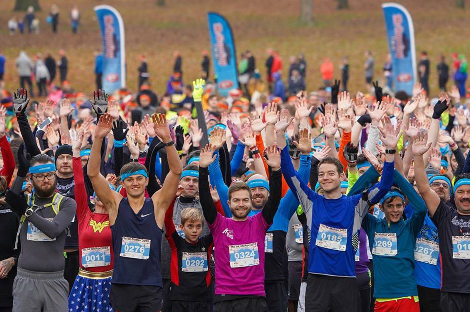 A large, enthusiastic group of runners wearing race bibs and costumes cheerfully raise their hands in the air before the start of a race. They stand outdoors on a grassy field with colorful banners in the background.