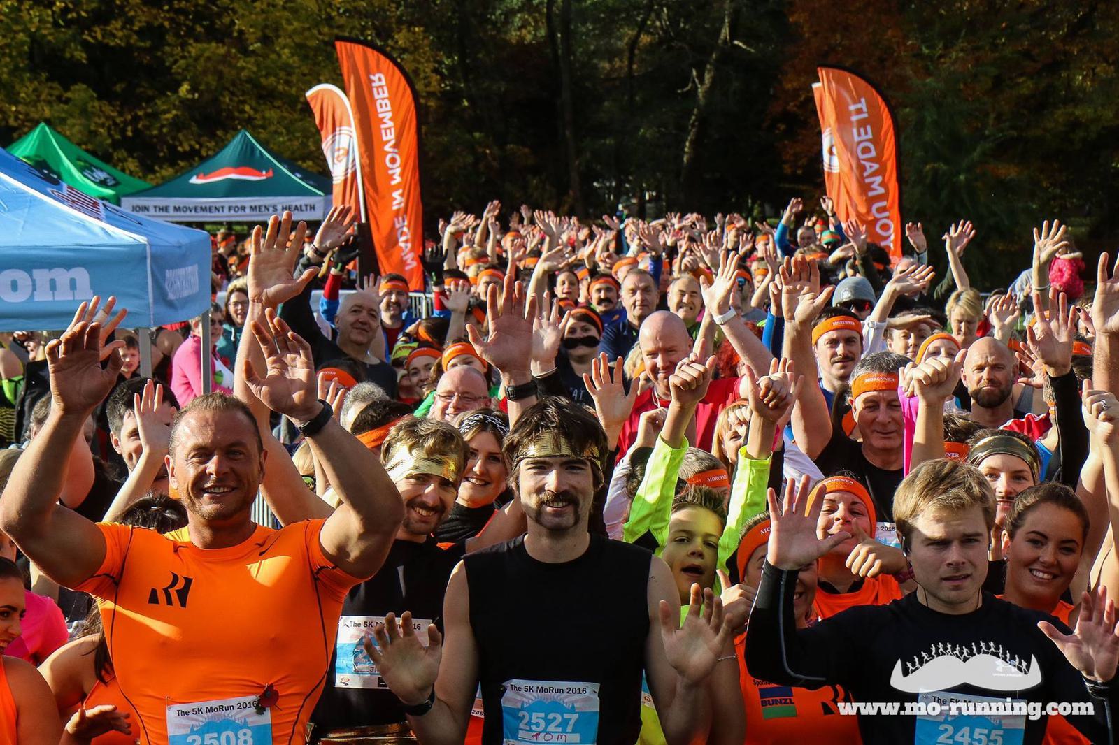 A large group of runners, many wearing orange shirts and headbands, gather at the starting line of a race. They are waving enthusiastically at the camera. Flags and tents are visible in the background. The atmosphere is energetic and excited.