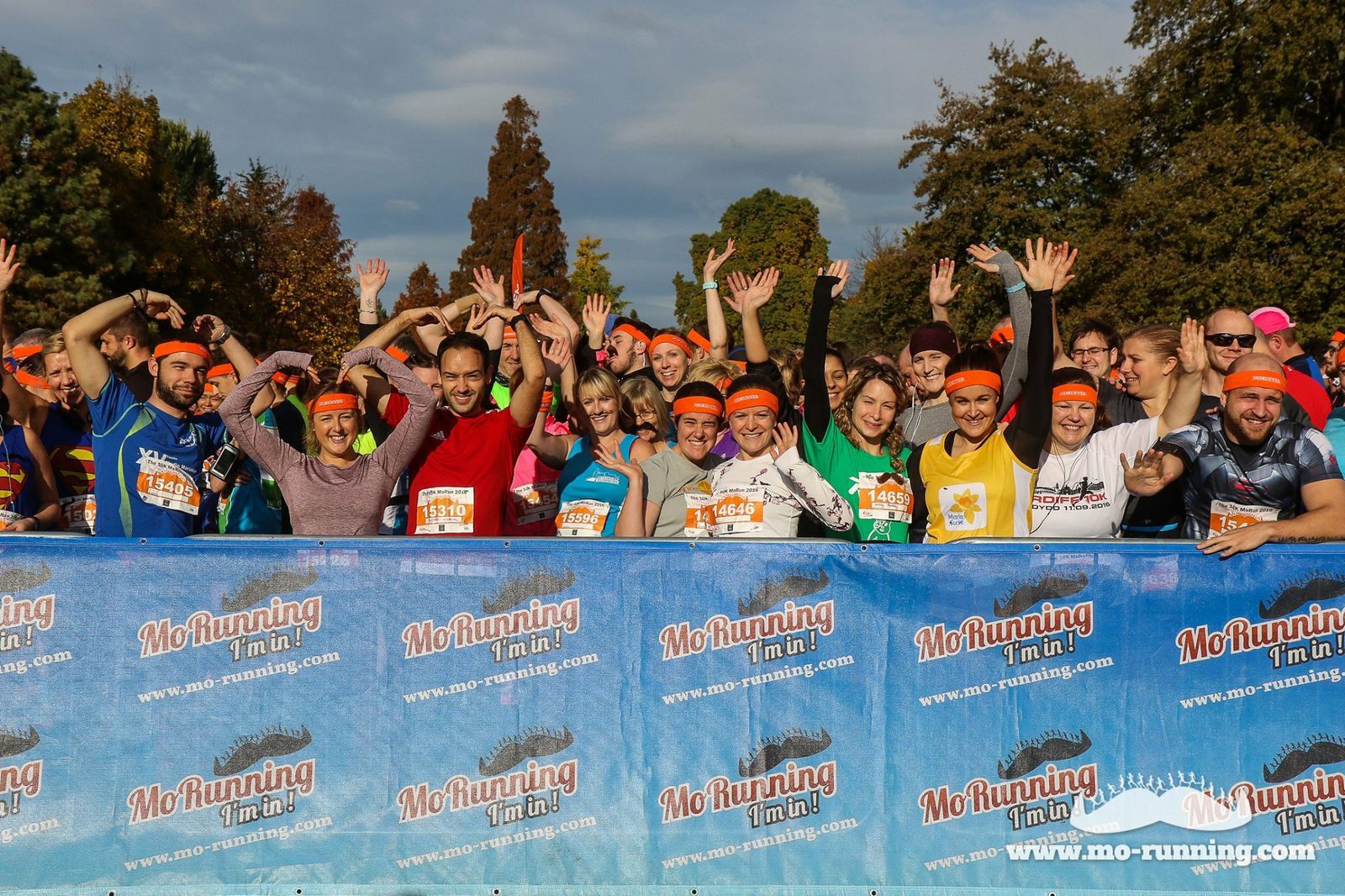 A diverse group of excited runners stand behind a blue banner with "MoRunning" printed on it. They are smiling, raising their arms, and wearing orange headbands with numbered race bibs. Trees and a partially cloudy sky are visible in the background.
