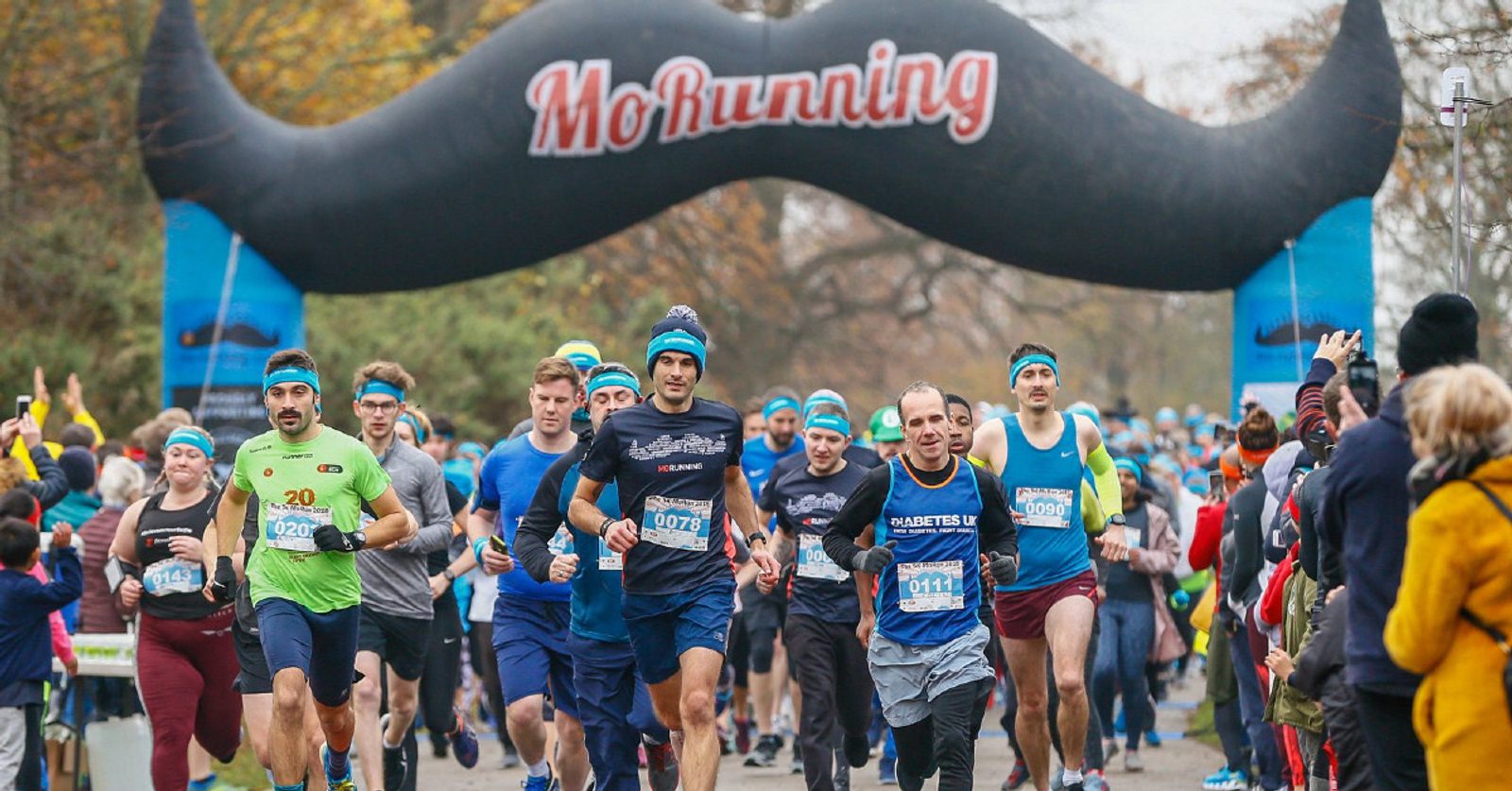 A large group of runners is participating in a race, passing under an inflatable arch marked with "MoRunning" and designed to look like a mustache. The runners wear numbered bibs and blue headbands, while spectators stand on the sidelines cheering them on.
