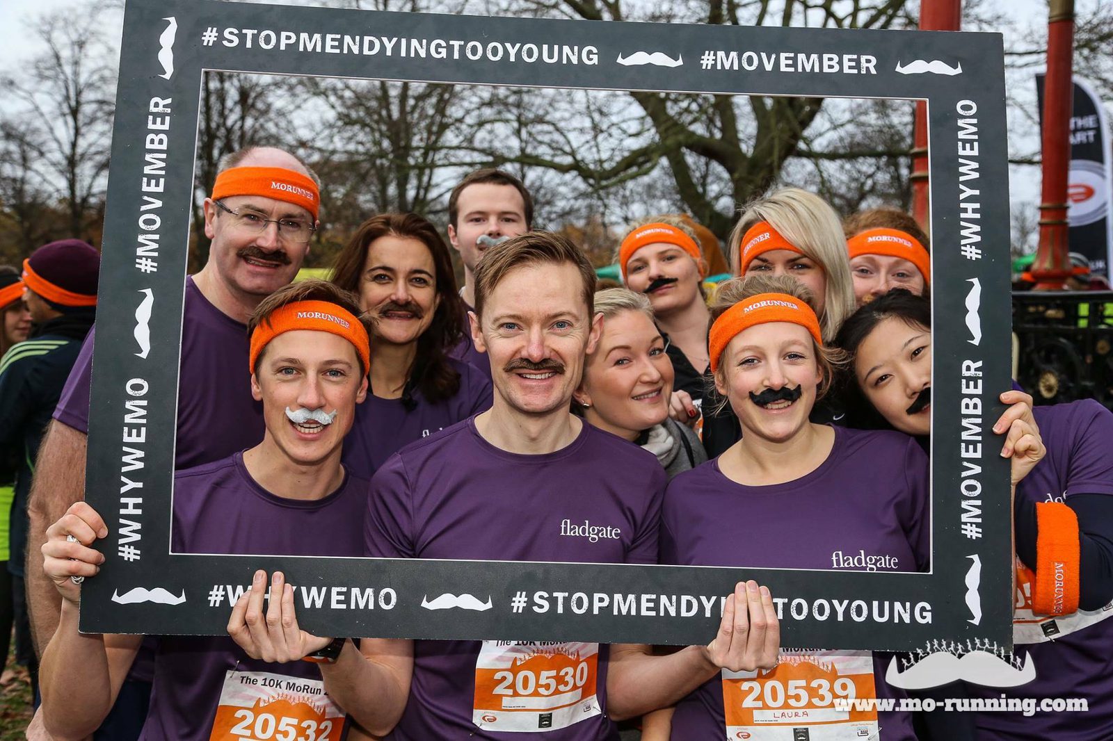 A group of people are posing together outdoors, all wearing purple shirts and orange headbands. Many have fake mustaches and they're holding up a large black frame with hashtags promoting Movember, including #STOPMENDYINGTOOYOUNG and #WHYWEMO.