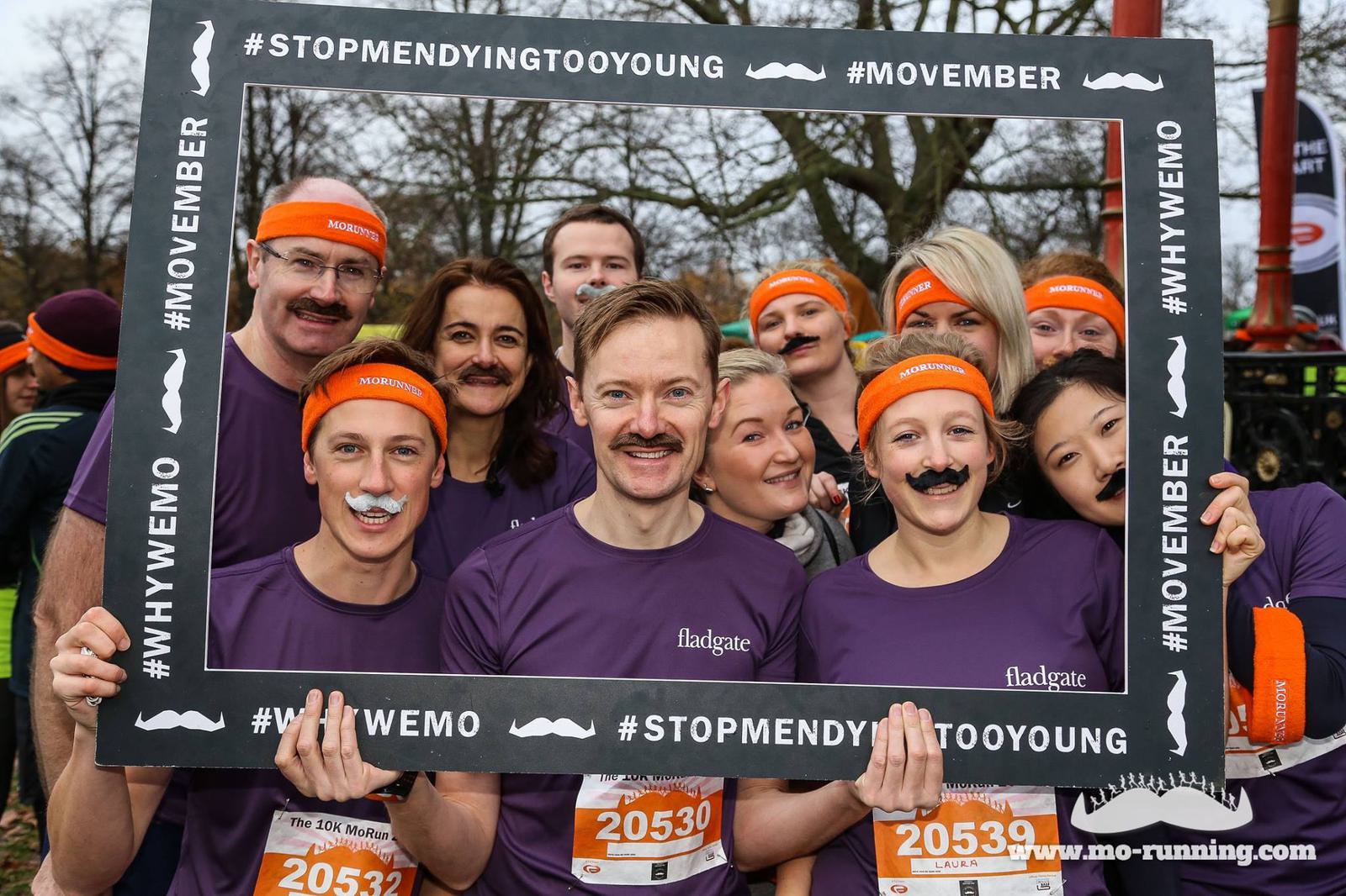 A group of people, wearing purple shirts and orange headbands, smile and pose for a photo while holding a large picture frame. They sport fake moustaches and wear race bibs. The frame reads "#STOPMENDYINGTOOYOUNG," "#MOVEMBER," and "#WHYWEMO.