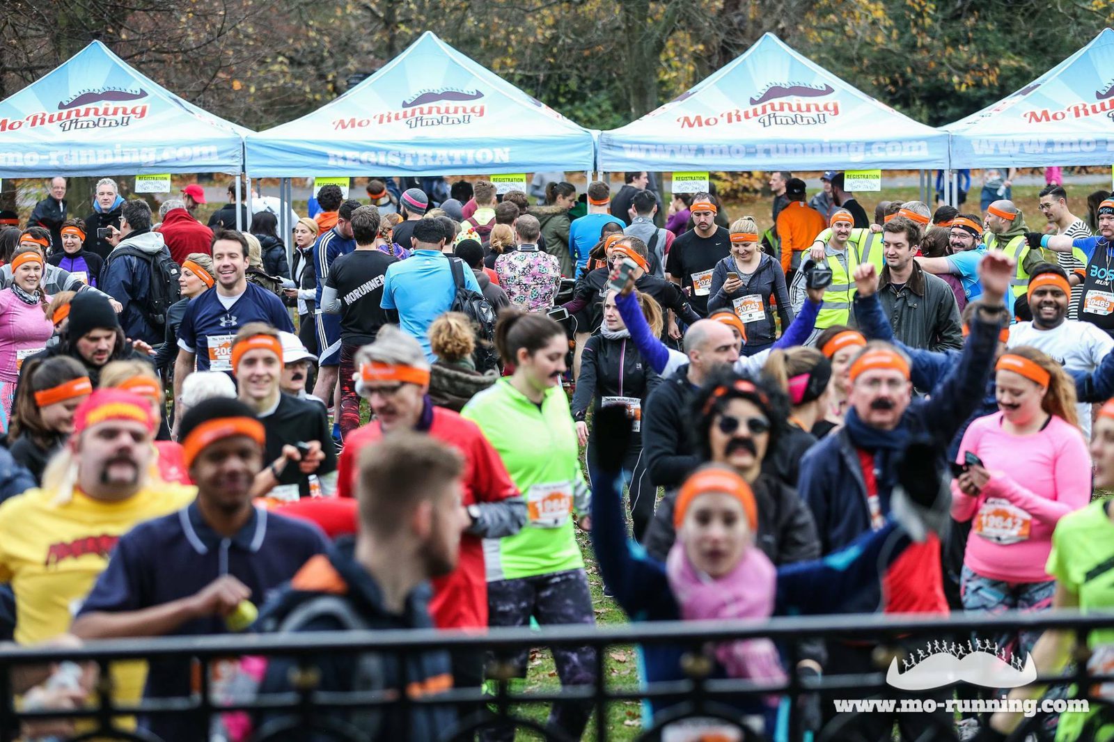 A crowd of people wearing colorful athletic clothing and orange headbands participate in an outdoor running event. Tents with event branding are visible in the background. Some participants are smiling, cheering, or taking photos. The event appears lively and energetic.