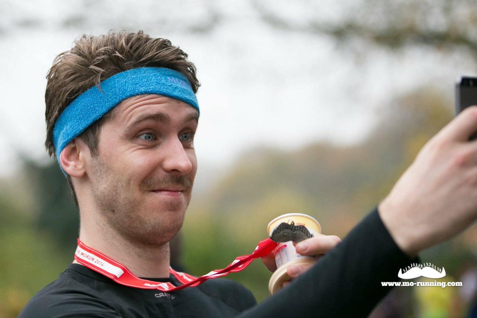 A person wearing a blue headband and a medal around their neck is holding what appears to be a small dessert cup. They are taking a selfie and have a playful expression on their face. The image includes the text "www.mo-running.com" in the bottom right corner.