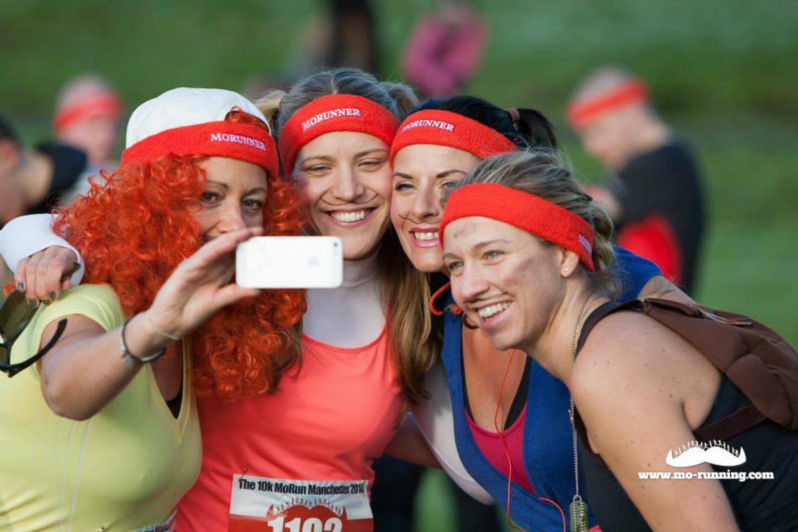 Four women wearing "MoRunner" headbands and sports attire are taking a selfie together during the 10k MoRun event. One woman has bright orange curly hair, possibly a wig. They are smiling and appear excited, with green grass and other participants in the background.
