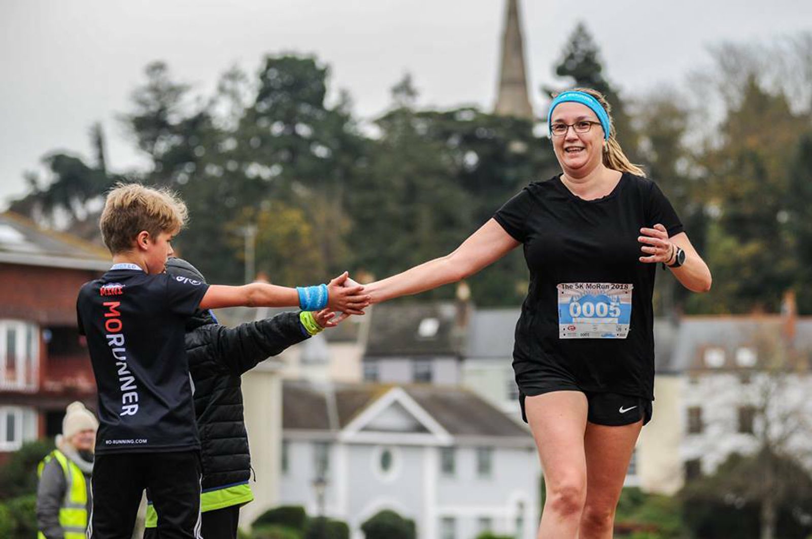 A female runner wearing a black outfit and a blue headband, with race bib 0005, reaches out to high-five a young boy in a "MORUNNER" shirt. The background features trees, houses, and a church steeple in a slightly blurred setting, indicating a running event.