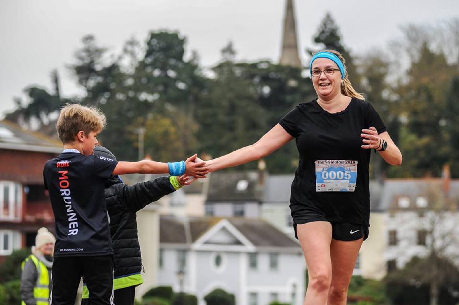 A runner wearing a black outfit and glasses high-fives a child with blond hair as she participates in a marathon. She is smiling and wearing a race bib, with buildings and trees in the background. Another person stands nearby, looking on.