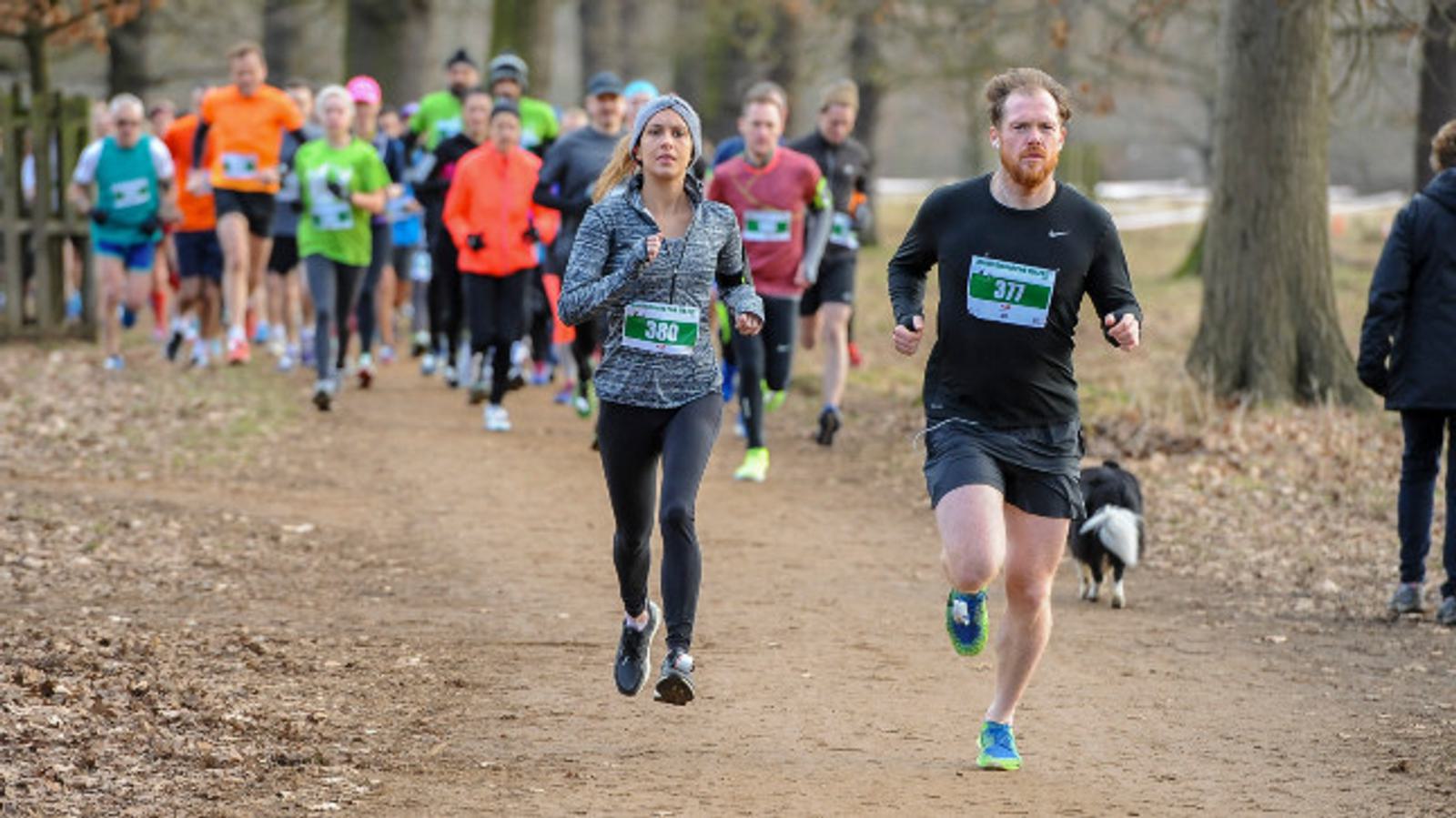 A man and a woman in athletic gear run on a dirt path surrounded by trees during a race. The man, wearing a black outfit, has a bib number 377, while the woman in black attire and a blue hat has 387. Other runners in colorful outfits follow behind them.