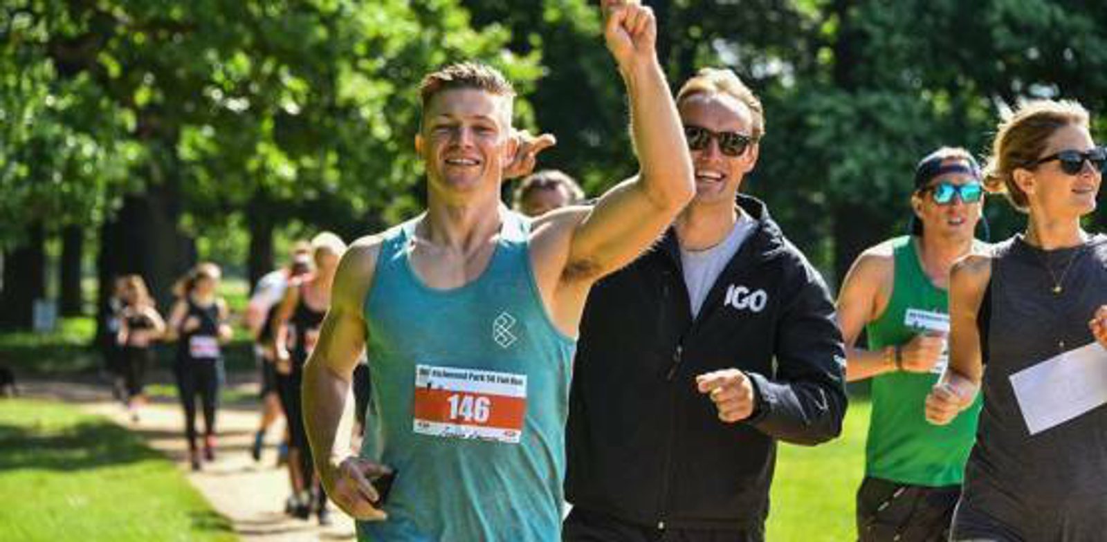 A group of runners outdoors, with a man in a green tank top and red race bib numbered 146 smiling and raising one arm. A man in sunglasses and a black hoodie beside him also smiles. Other runners in casual athletic wear are in the background on a sunny day.
