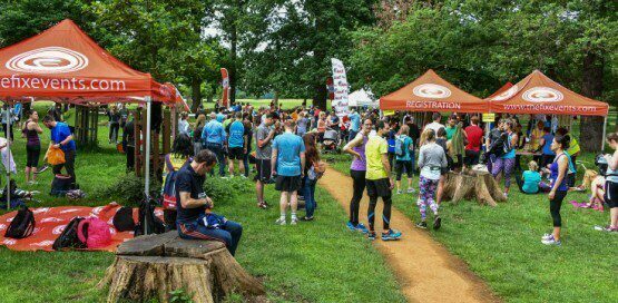A large group of people gathers in an outdoor area for a running event. There are two orange tents labeled "enfxevents.com" and "REGISTRATION." Participants are seen talking, preparing, and relaxing on tree stumps. The background shows trees and a grassy area.