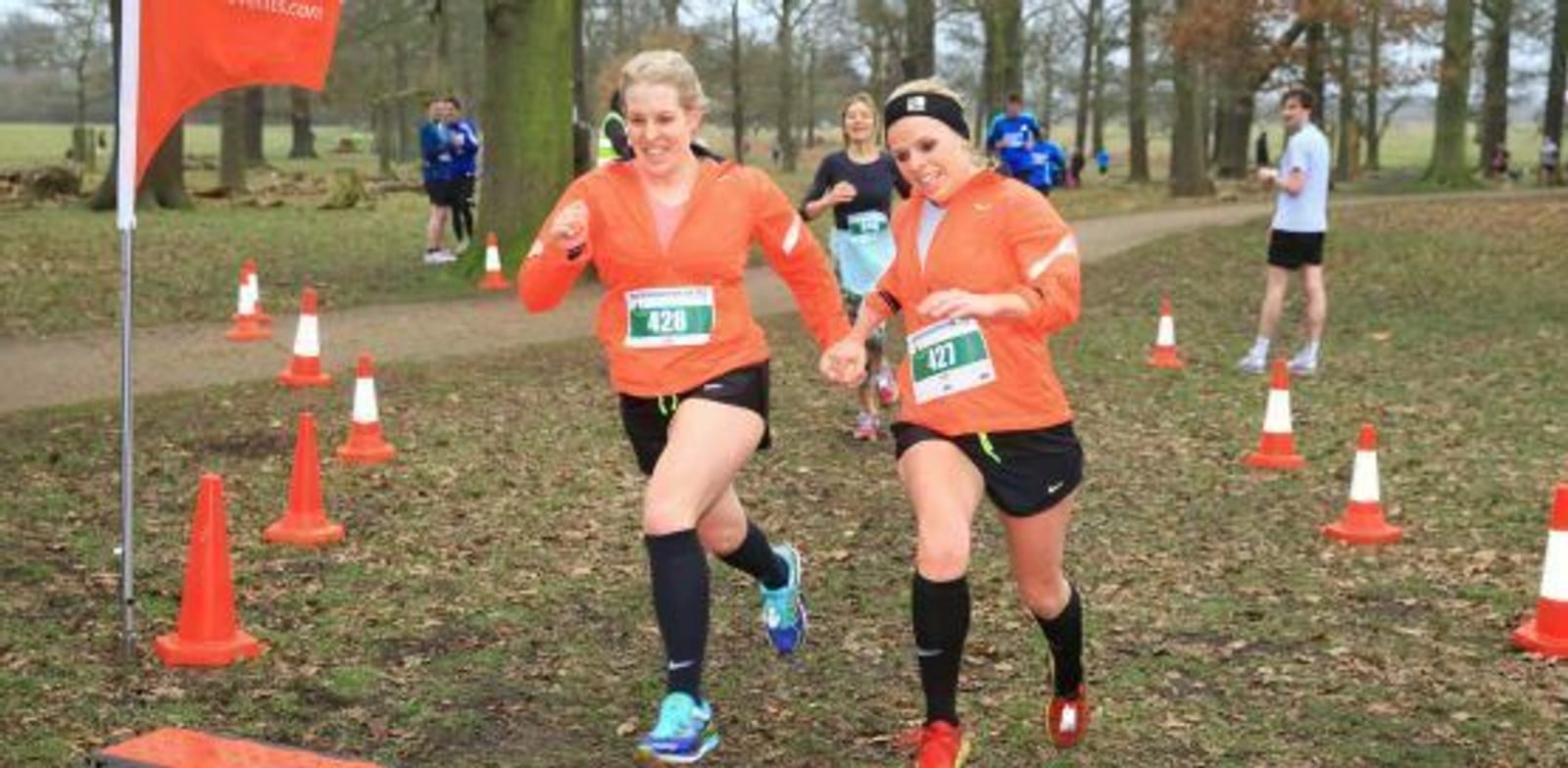 Two runners wearing matching orange shirts and black shorts are approaching the finish line of a race, each with a race bib pinned to their shirt. They are surrounded by orange cones and trees. Other participants are seen in the background.