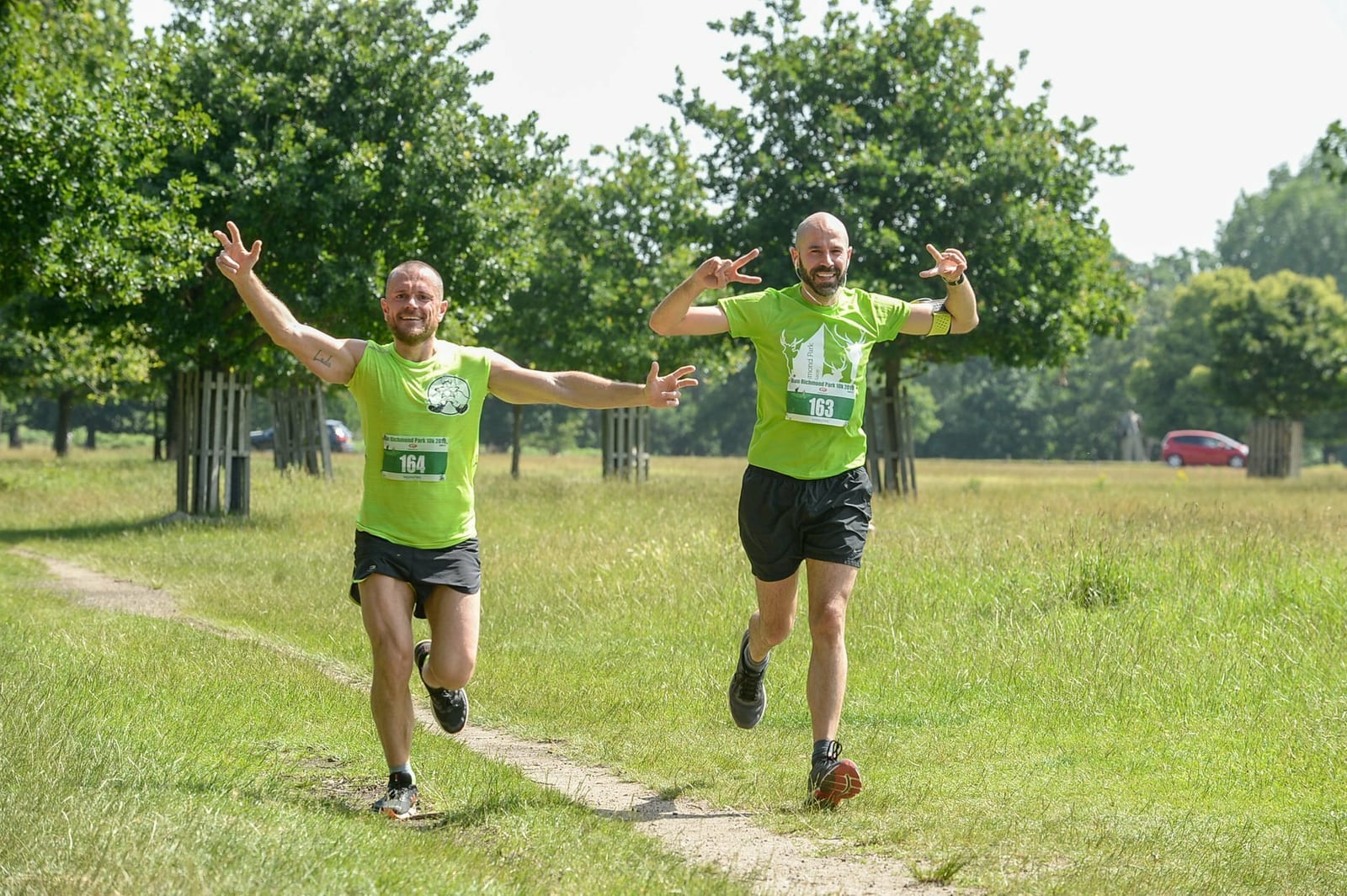 Two men in green sports shirts and black shorts are running on a trail in a park. Both have bib numbers on their chests. One has "164" and the other has "162". They are smiling and raising their arms with a backdrop of trees and grass on a sunny day.