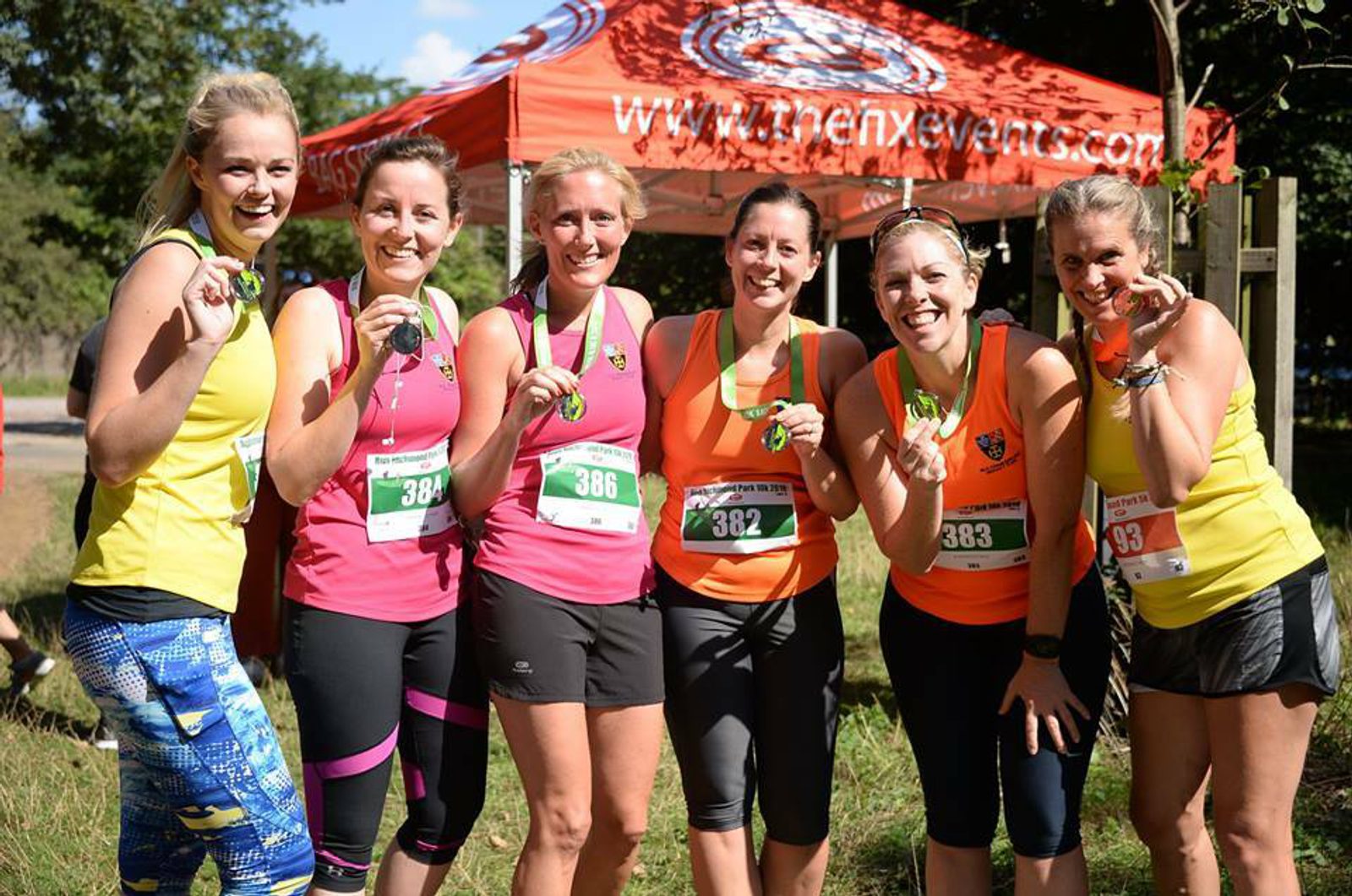 Six smiling women wearing colorful athletic clothing and holding medals, standing in front of a red tent with the logo "thenxevents" during a sunny outdoor event. They have race bibs containing different numbers pinned to their outfits.