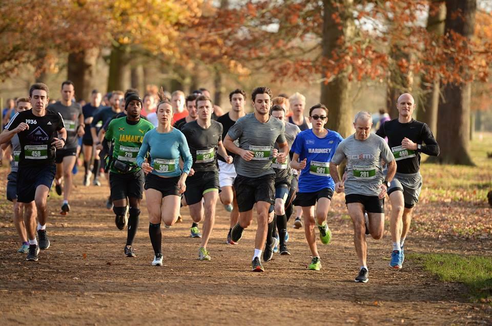 A large group of runners, both men and women, are participating in a race on a dirt path in a park during autumn. They are surrounded by trees with golden leaves. The runners are wearing various athletic outfits and race bibs. They appear focused and determined.