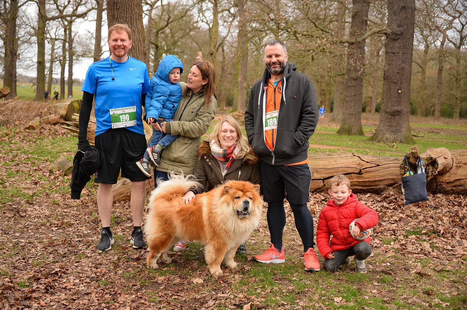 A group photo in a park features two men, two women, and two children dressed in running gear and casual clothes. The adults wear race bibs, with one carrying a child. A fluffy dog stands in the center. The background shows trees and fallen leaves.