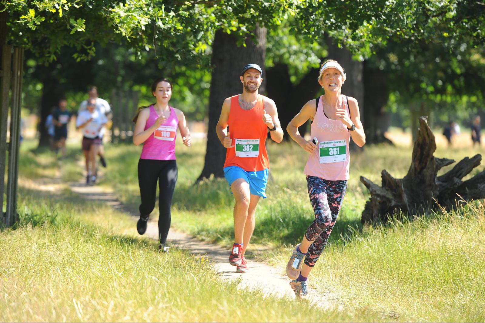 Three runners participating in a race outdoors on a dirt path, surrounded by lush greenery. The woman on the right wears a pink tank top and patterned leggings, the man in the center sports a red tank top and blue shorts, and the woman on the left wears a purple tank top and black leggings. They all have bib numbers pinned to their shirts.