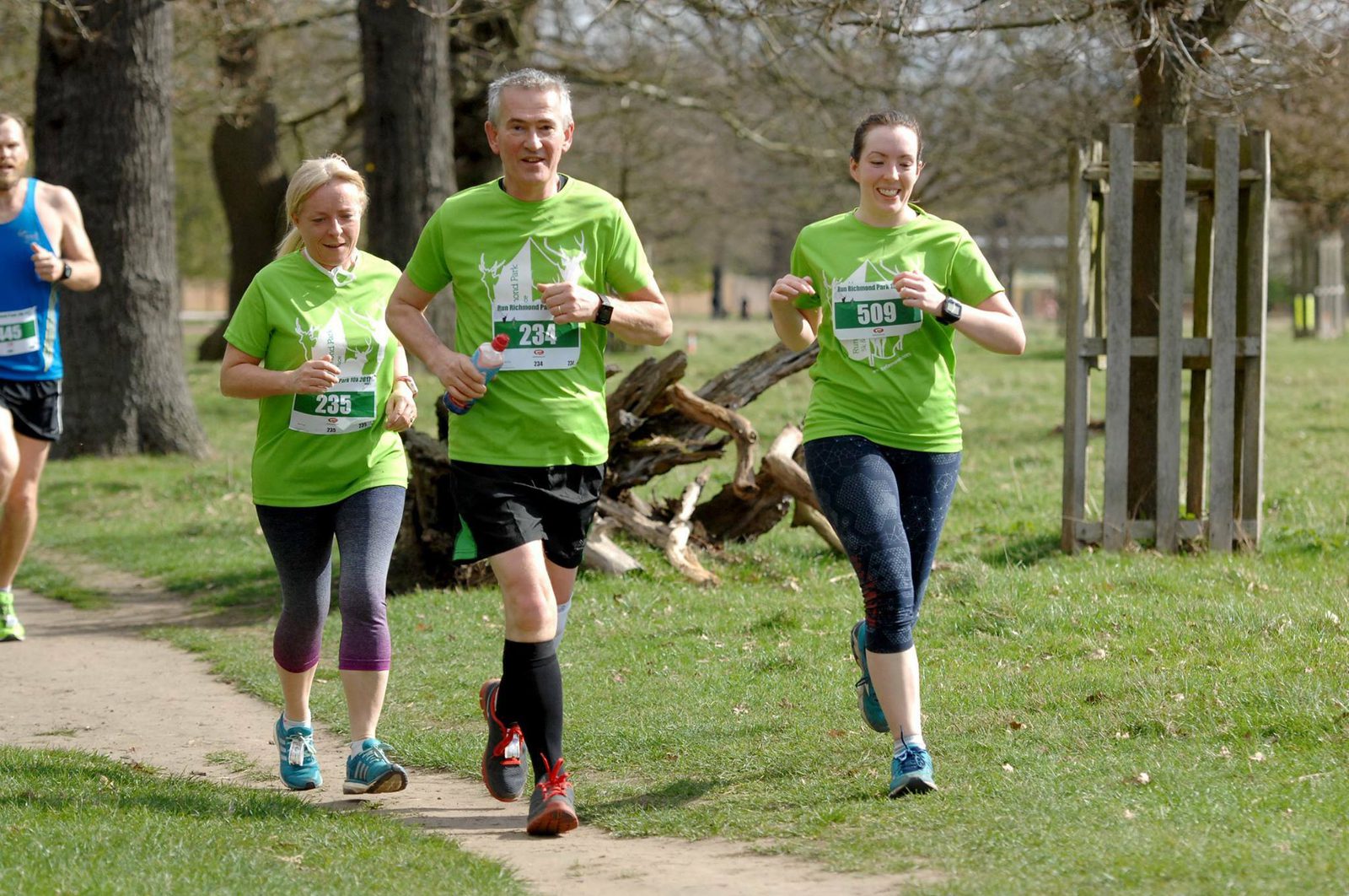 Three runners wearing matching green t-shirts are participating in an outdoor race on a dirt path lined with grass and trees. The man in the center is flanked by two women. They appear to be enjoying the event, running closely together and smiling.